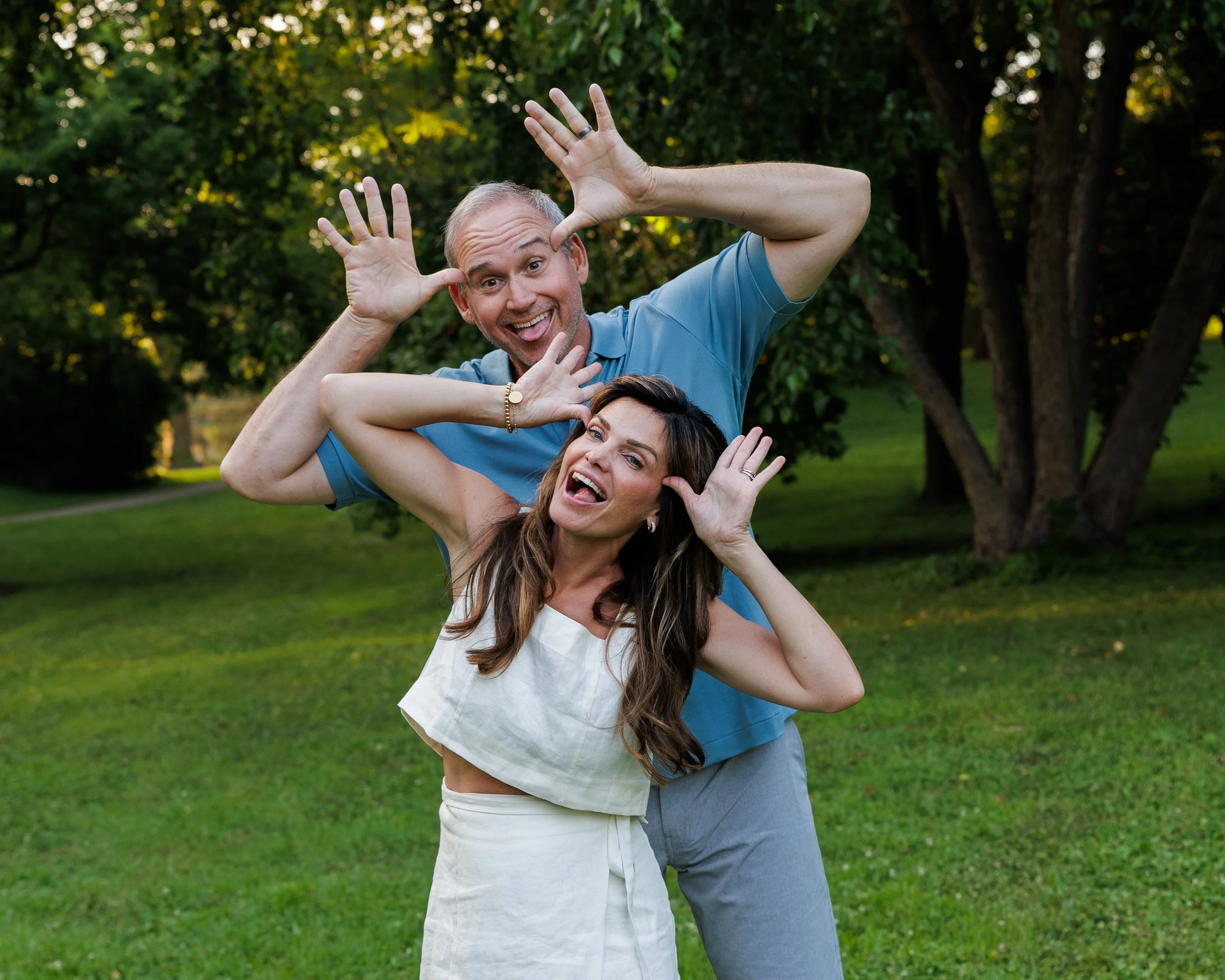 A man and woman making playful faces outdoors in a park with trees and grass.