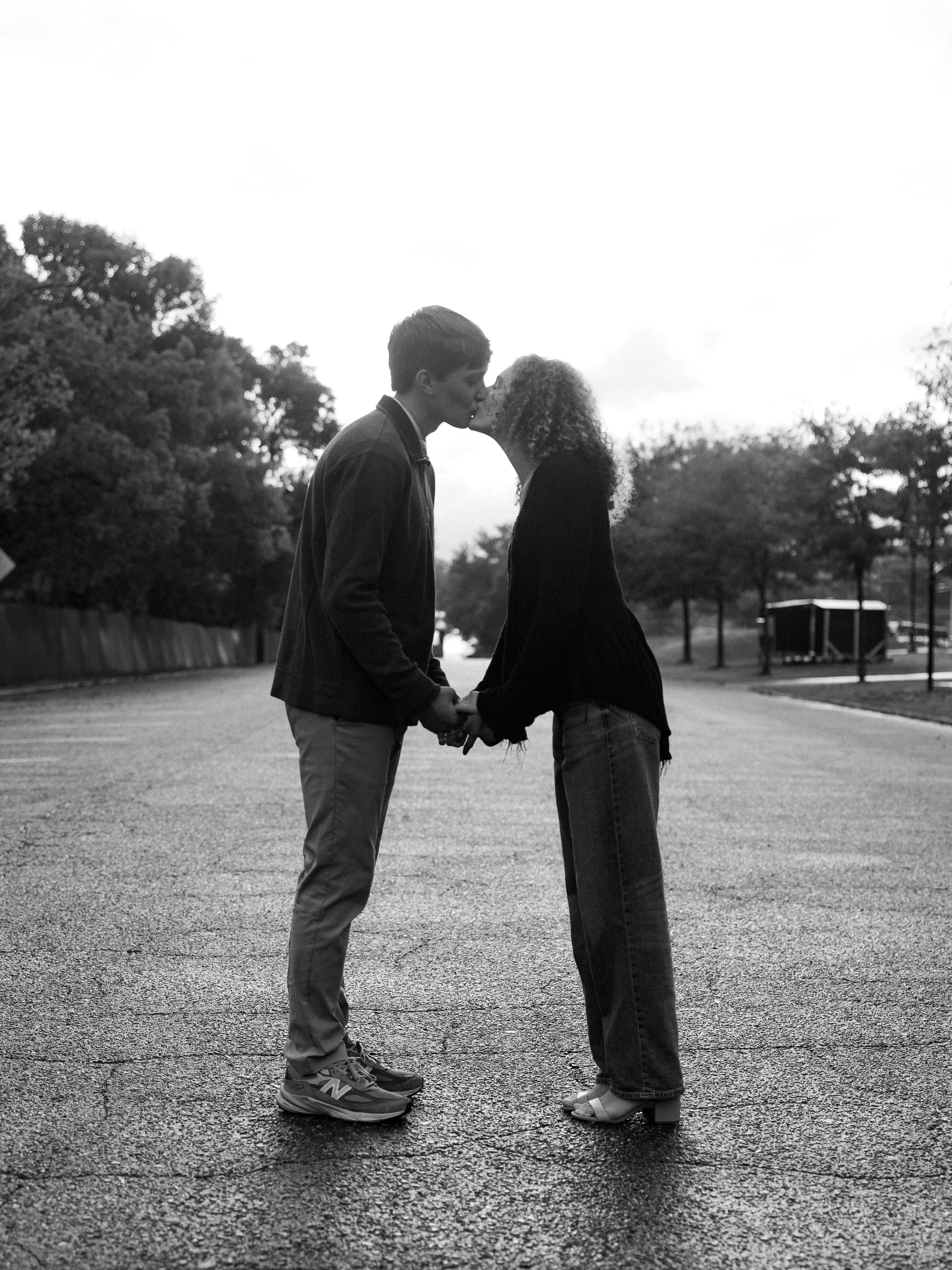 A black and white photo of a young couple kissing and holding hands on an empty street, with trees and a fence in the background.