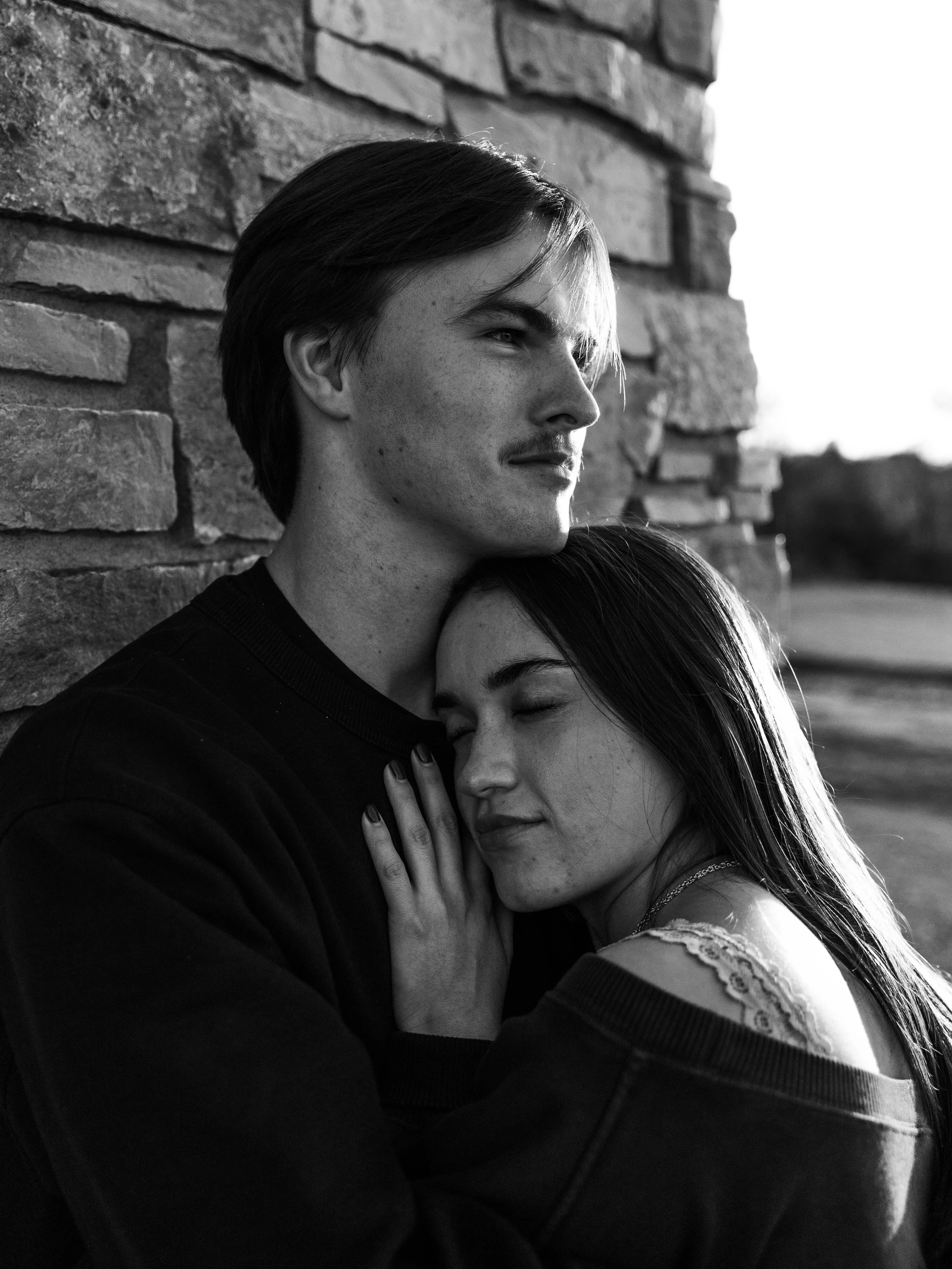 A black and white photo of a young couple, a man and woman, leaning against a brick wall. The woman has her eyes closed and is hugging the man, who is looking into the distance.