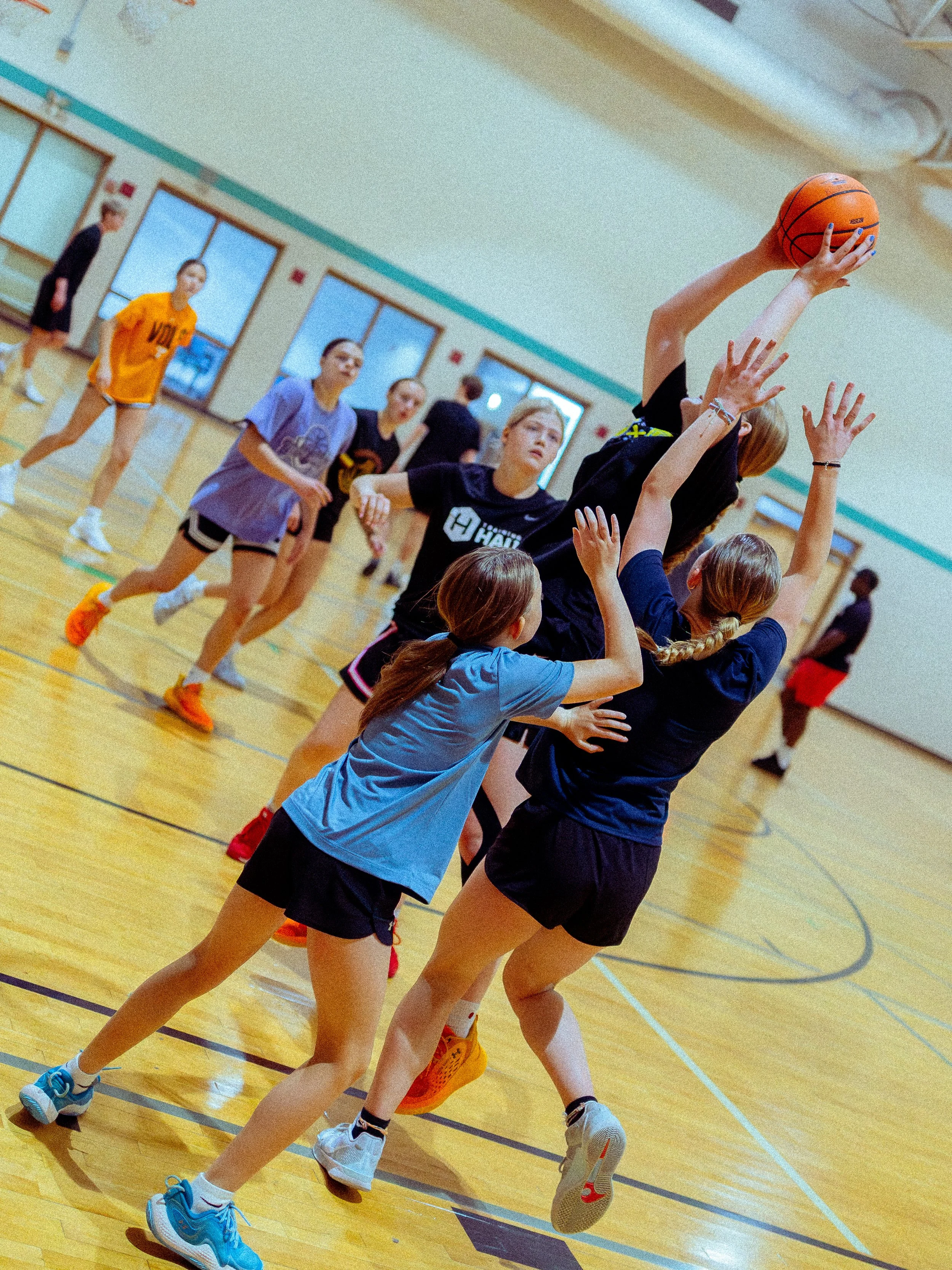 Young girls playing basketball indoors, one girl jumping to shoot or block the ball while others defend or reach for it.