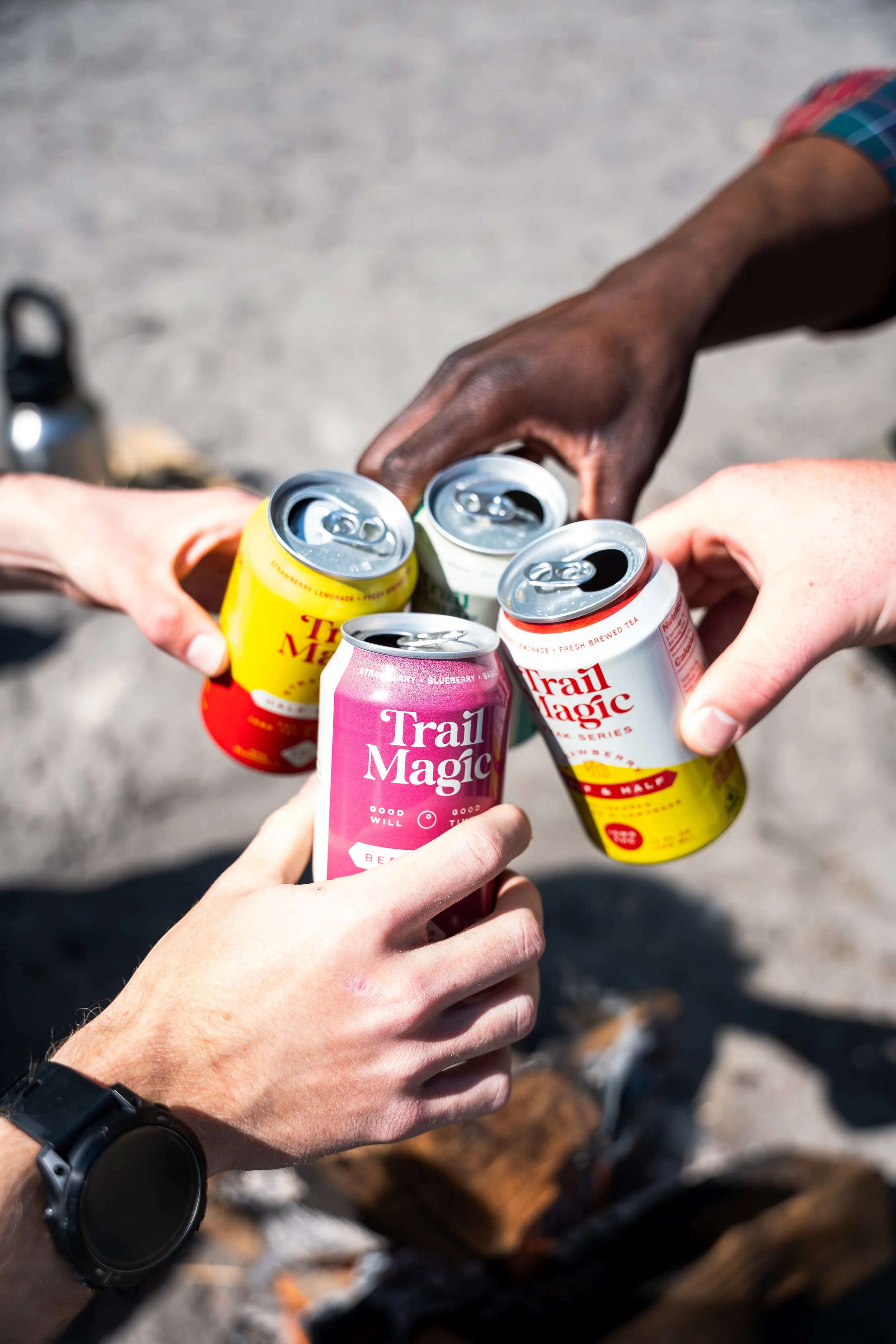 Four cans of Trail Magic drinks being clinked together in a toast outdoors on a sandy ground.