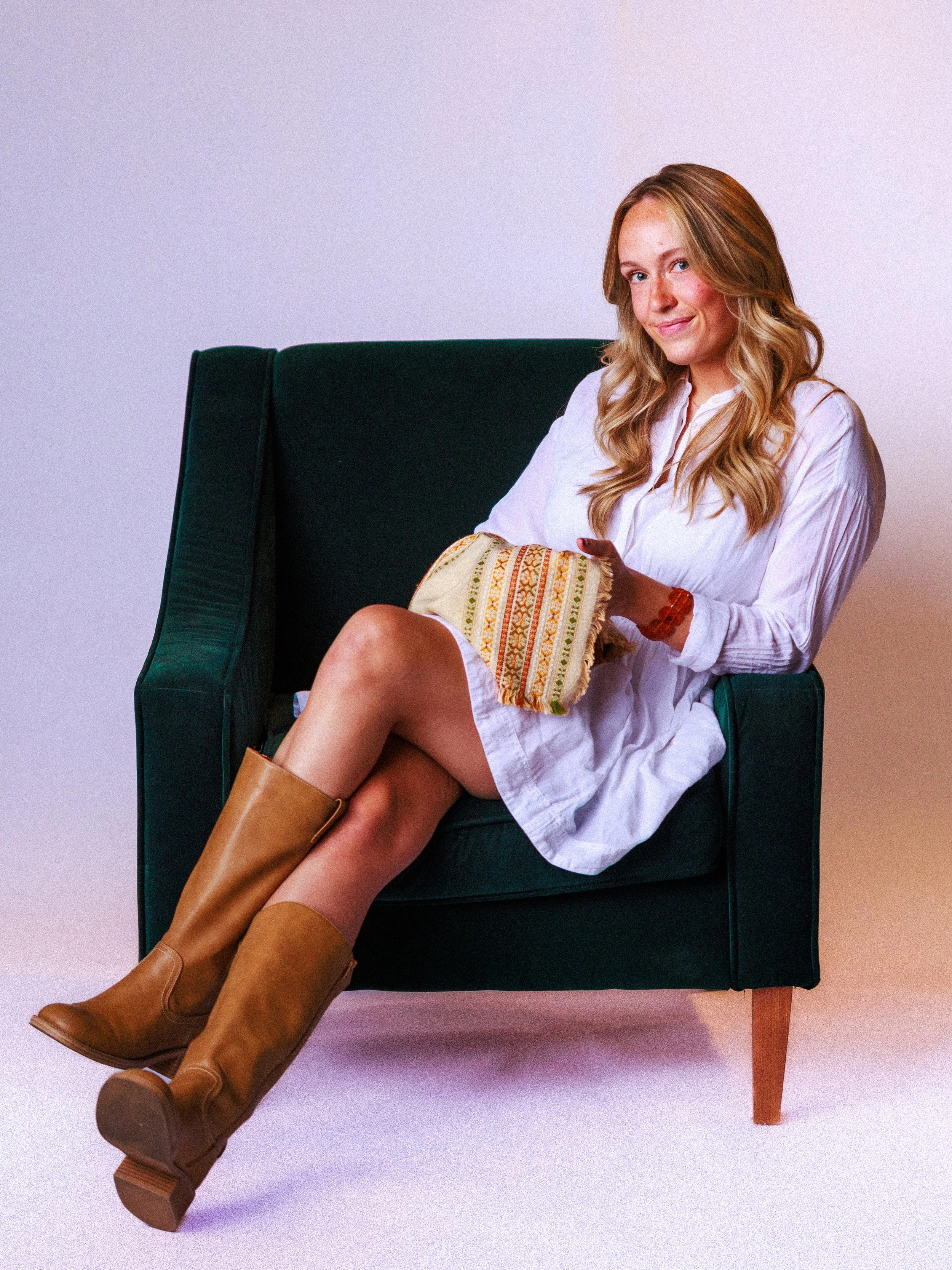 A woman with long blonde hair sitting on a dark green armchair, wearing a white dress, brown knee-high boots, and holding a colorful cloth. She is smiling and looking at the camera against a plain light background.