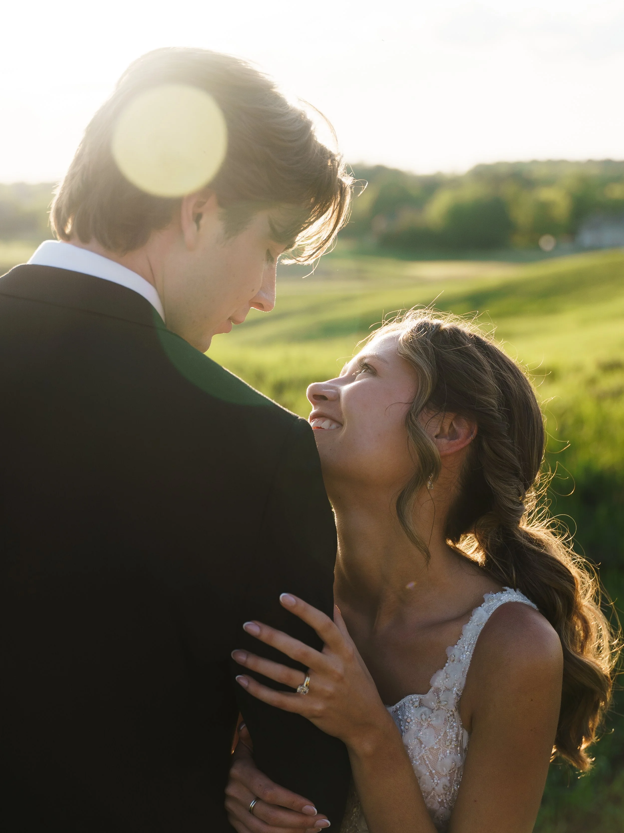 A romantic moment between a bride and groom outdoors during sunset, embracing and looking into each other's eyes with a green landscape in the background.