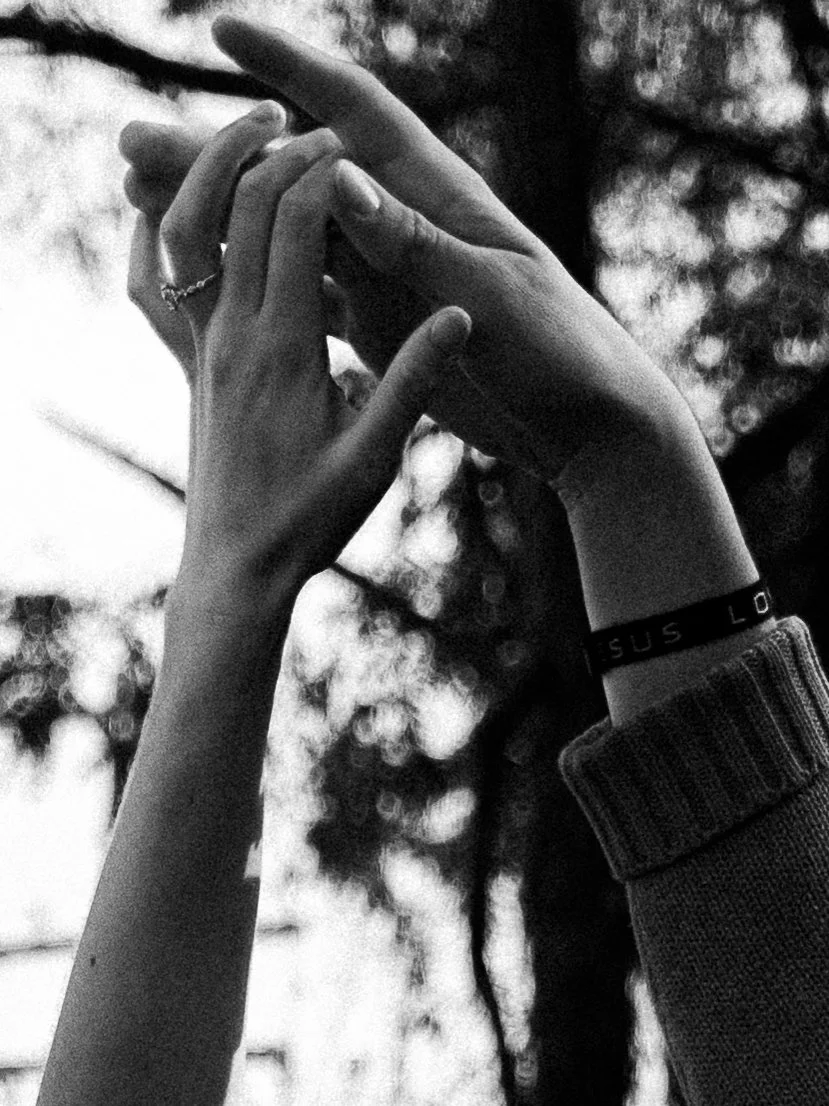 Black and white photo of two hands gently touching, with one hand wearing a ring and a wristband, and the background shows blurred trees.