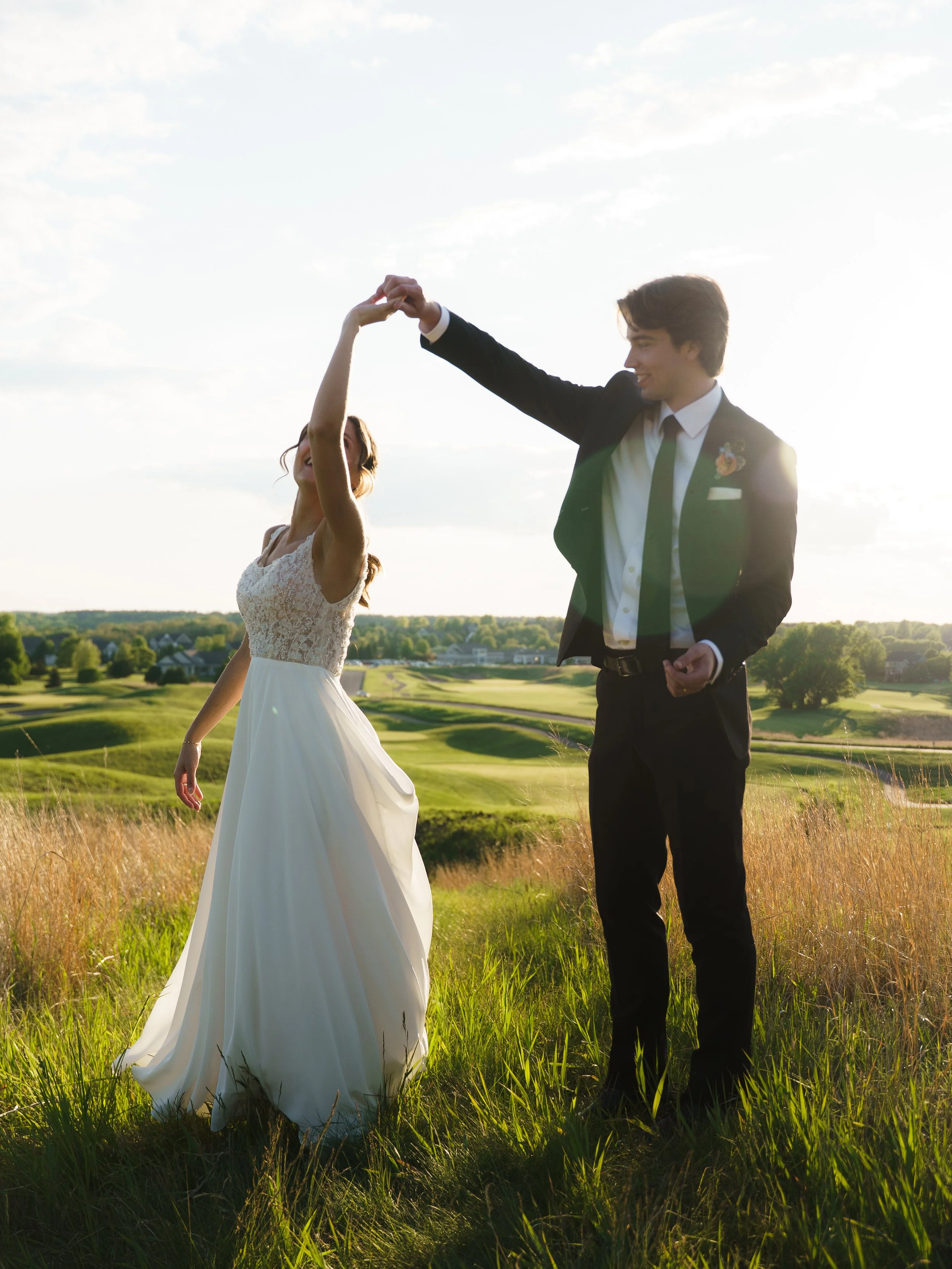 A bride and groom dancing outdoors on a grassy field at sunset.