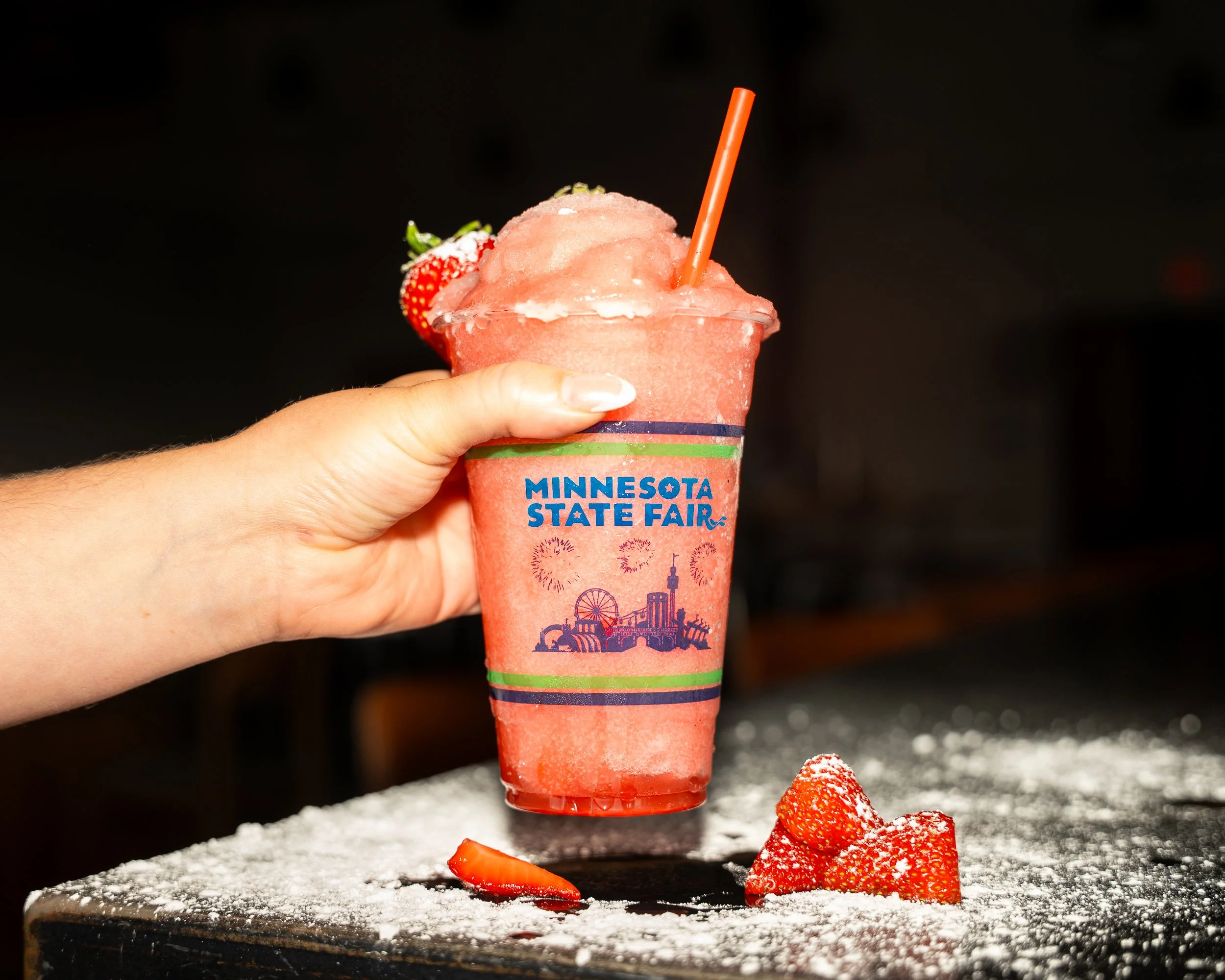 A hand holding a pink strawberry smoothie in a cup with a straw, labeled 'Minnesota State Fair,' with a strawberry garnish, against a dark background, on a surface dusted with powdered sugar and strawberry slices.