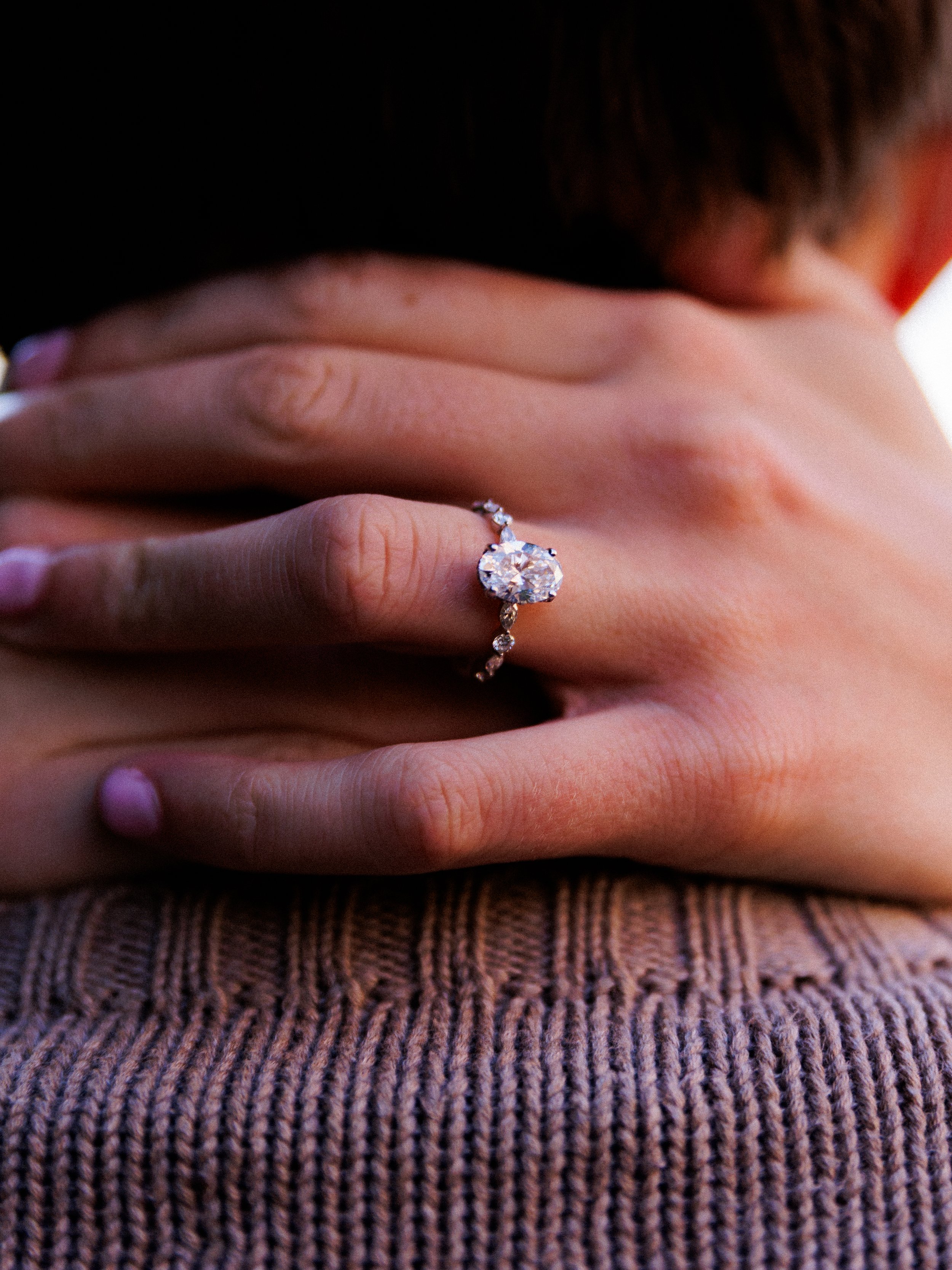 Close-up of a woman's hand wearing an engagement ring with a large oval diamond surrounded by smaller diamonds on a delicate band.