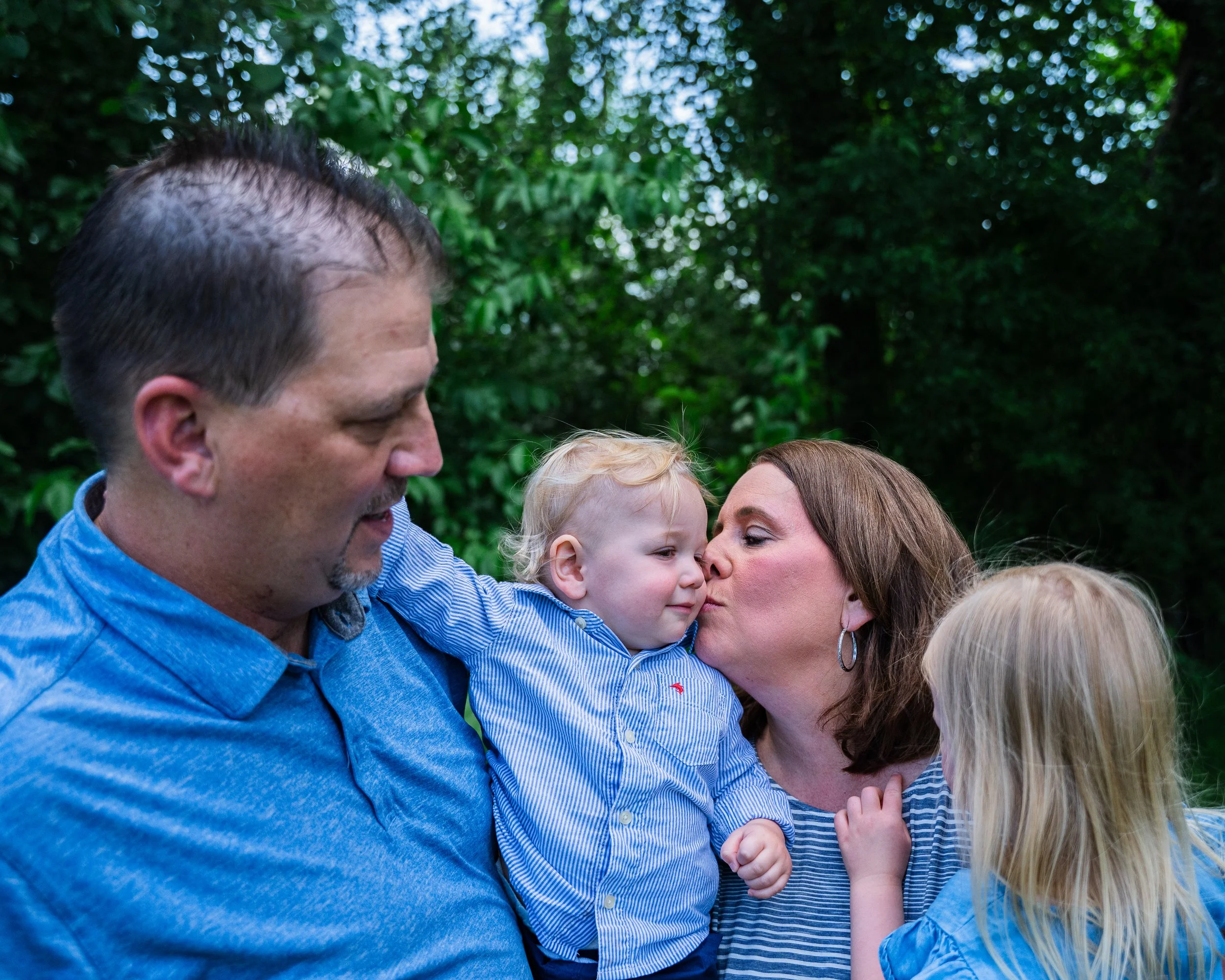 A family of four outdoors with greenery in the background. The father is holding a young boy, the mother is giving a kiss to the boy, and a young girl is touching the mother's shoulder.