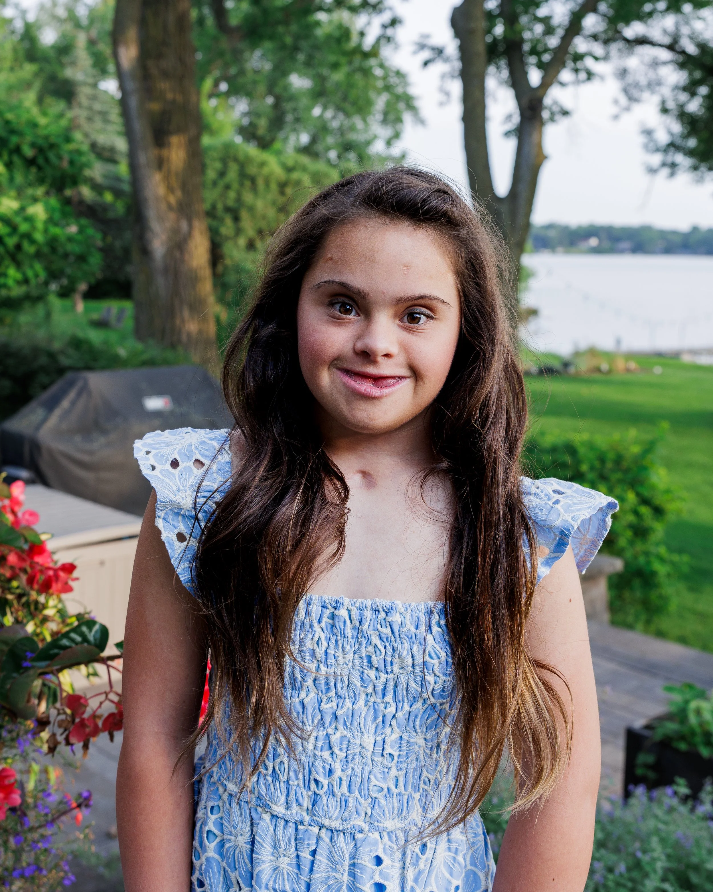A young girl with long brown hair, wearing a blue dress with ruffled sleeves, standing outdoors near a lake with trees and greenery in the background.