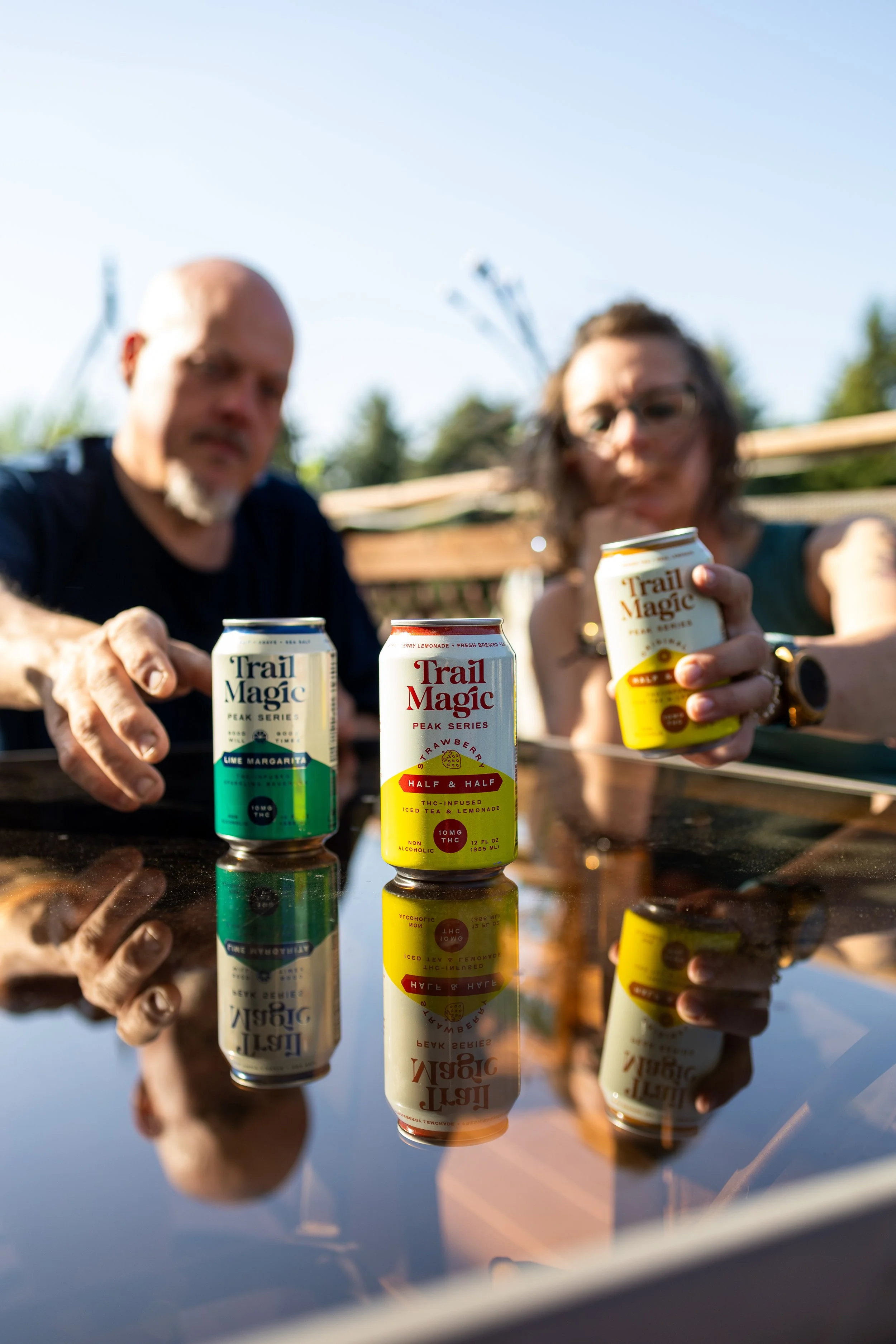 Two people holding cans of Trail Magic beverages, with one can in focus on a reflective glass surface, featuring cans labeled Lime Margarita, Strawberry, and Apple Cider, outdoors on a sunny day.