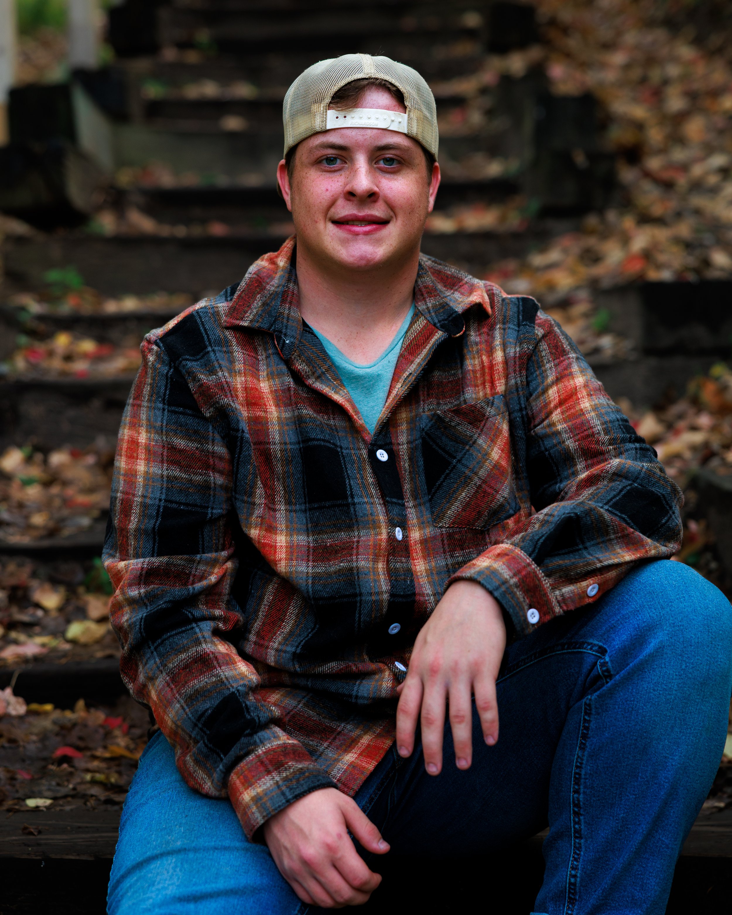 Young man sitting outdoors on wooden stairs covered with fallen leaves, wearing a plaid shirt and a backwards baseball cap.