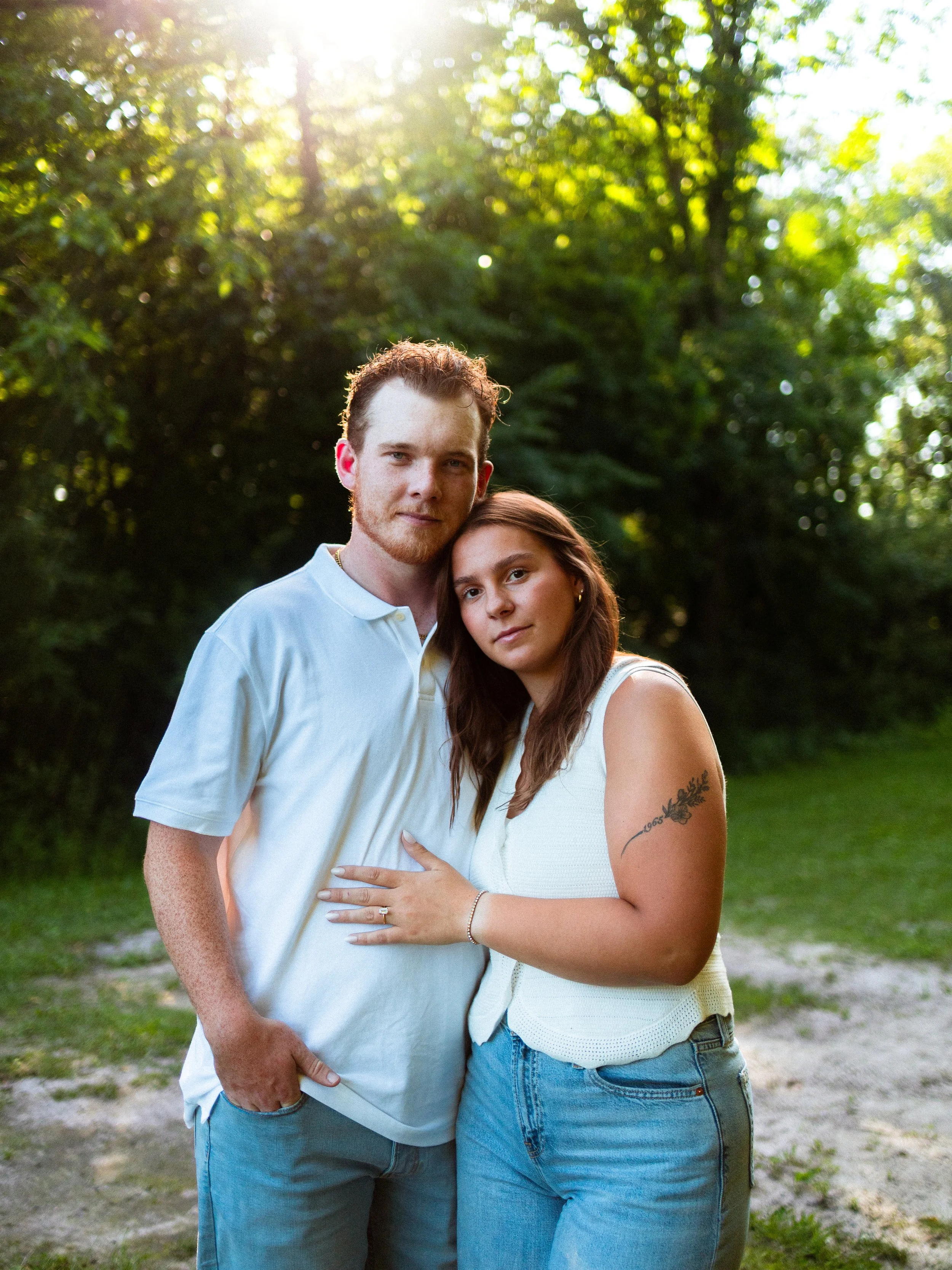 A young couple standing close together outdoors, with sunlight filtering through green trees in the background. The man is wearing a white polo shirt, and the woman has long brown hair, wearing a sleeveless white top, and has a tattoo on her upper ar