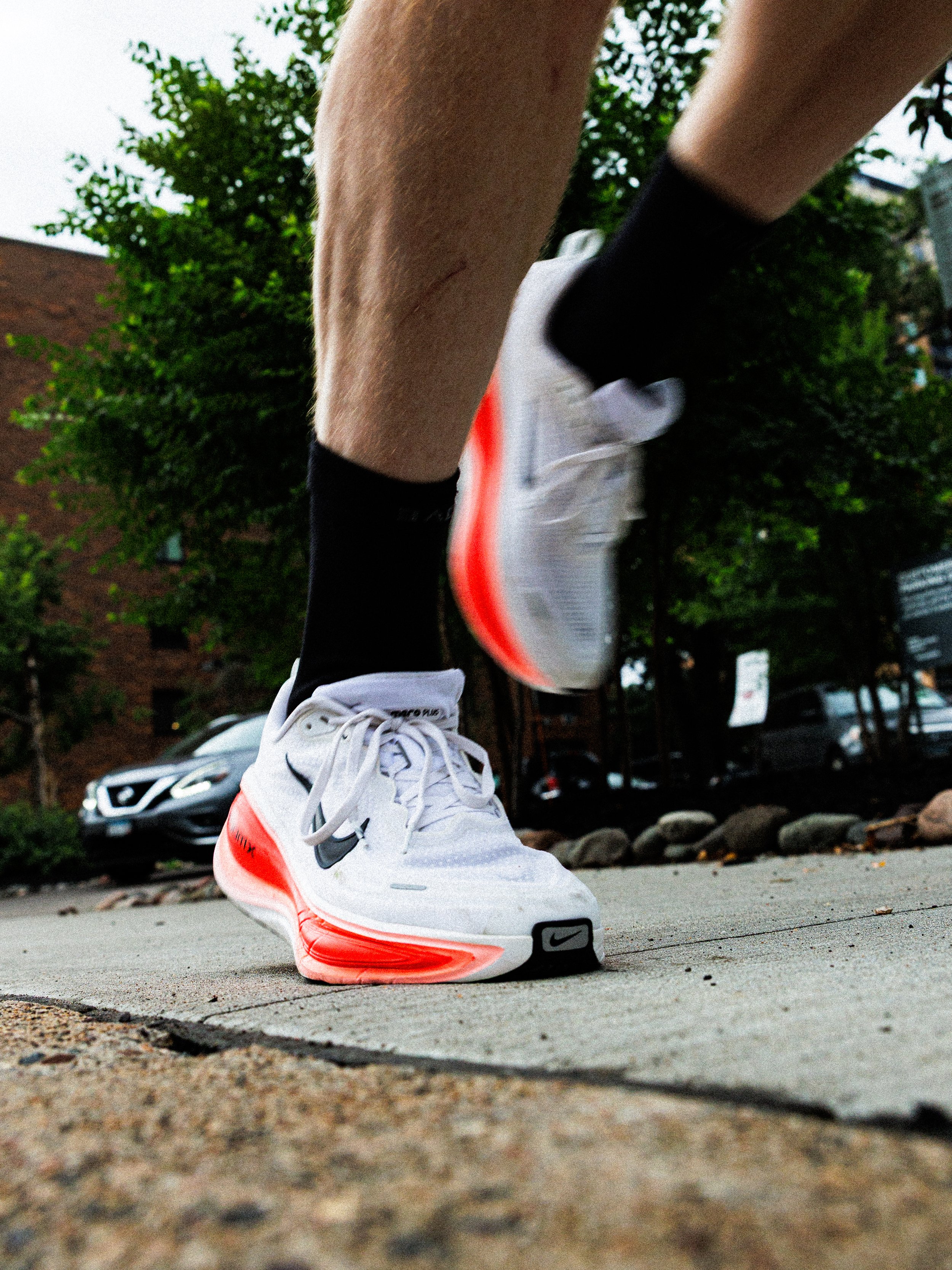 Close-up of a person's legs wearing running shoes and black socks, preparing to start running on a sidewalk with trees and parked cars in the background.