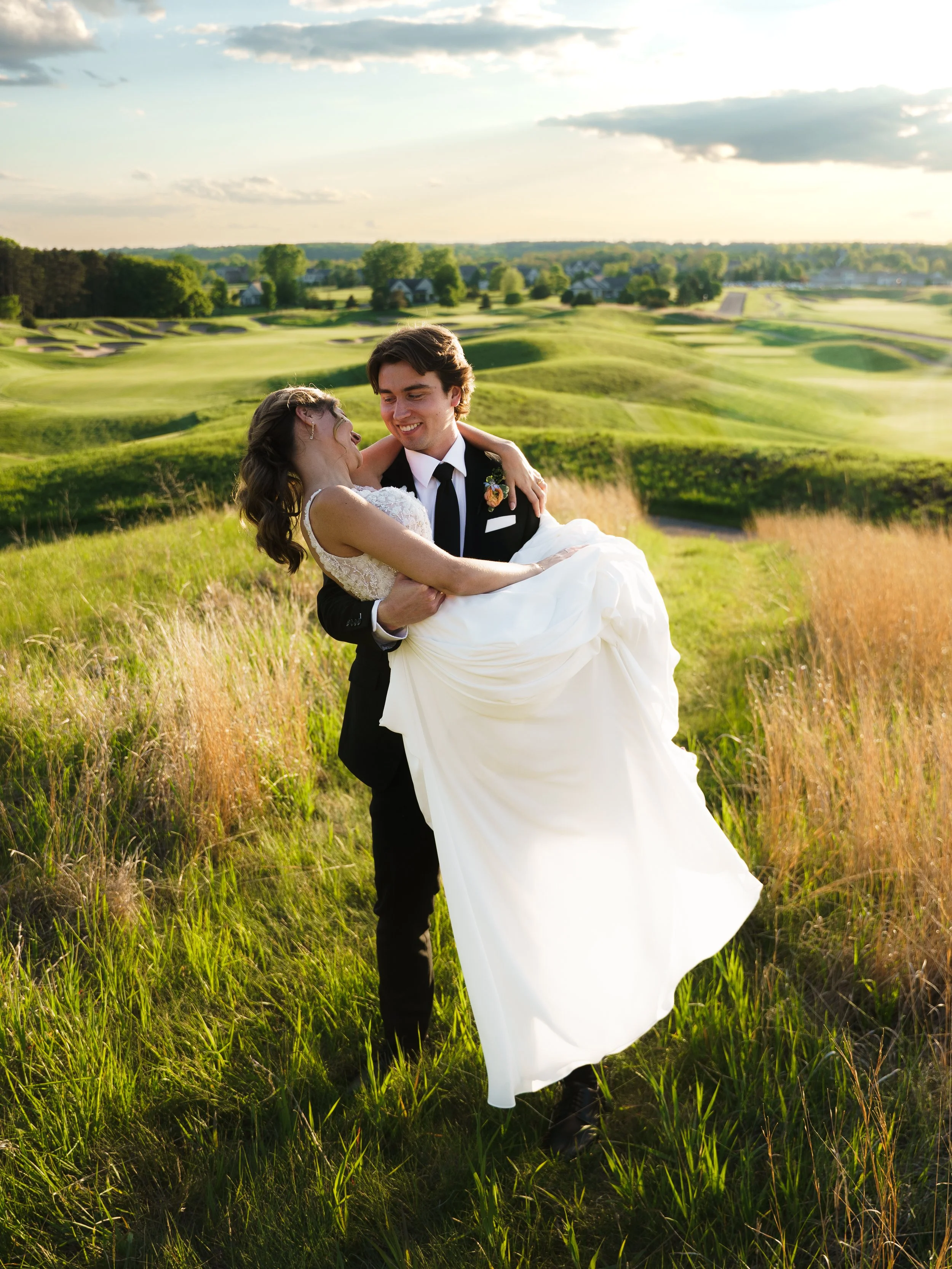 A young man in a tuxedo holding a smiling woman in a wedding dress in a grassy field with a golf course landscape and houses in the background, during sunset.