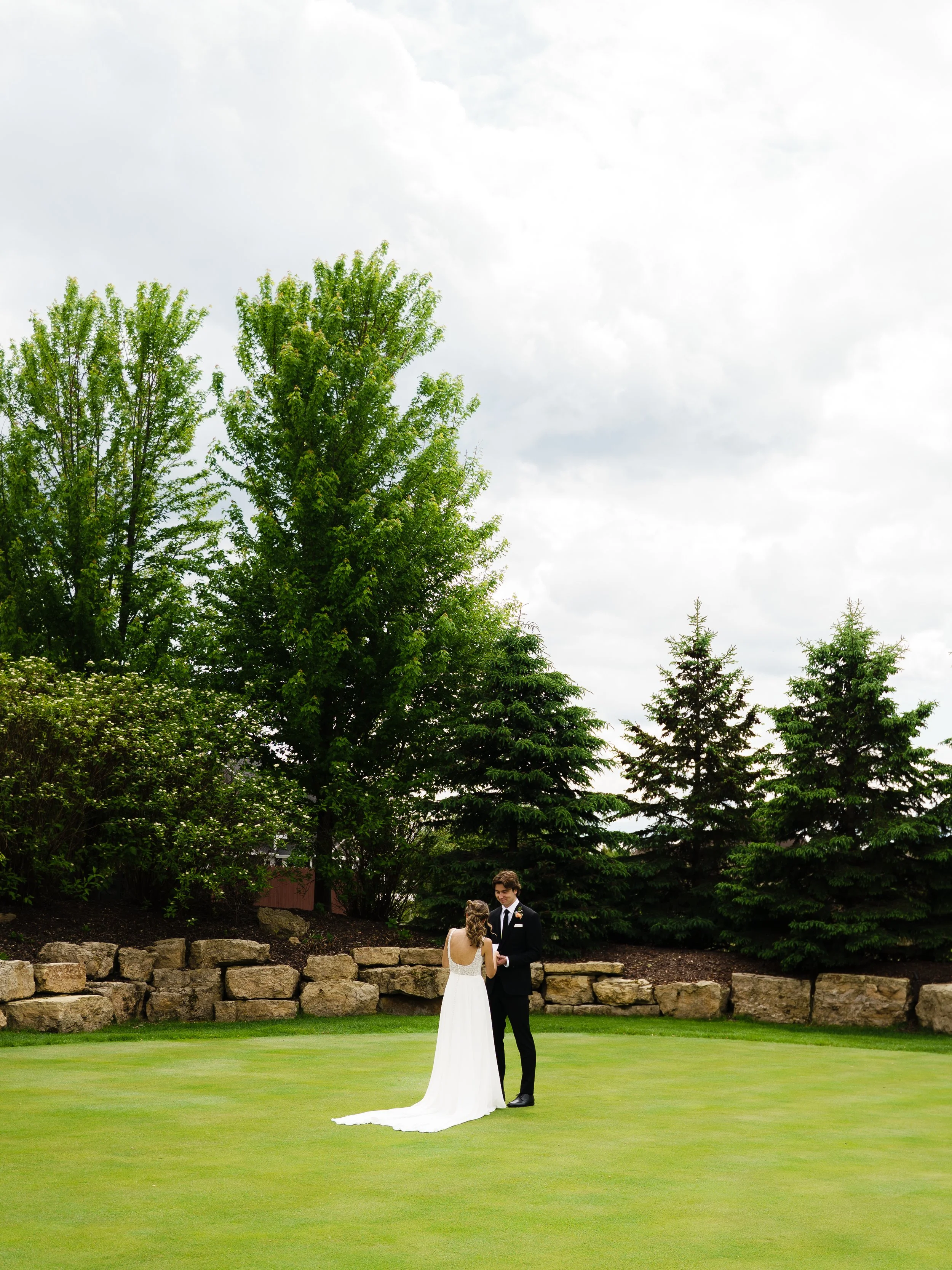 A couple dressed in wedding attire standing on a grassy area surrounded by trees and rocks, exchanging vows or holding hands outdoors.