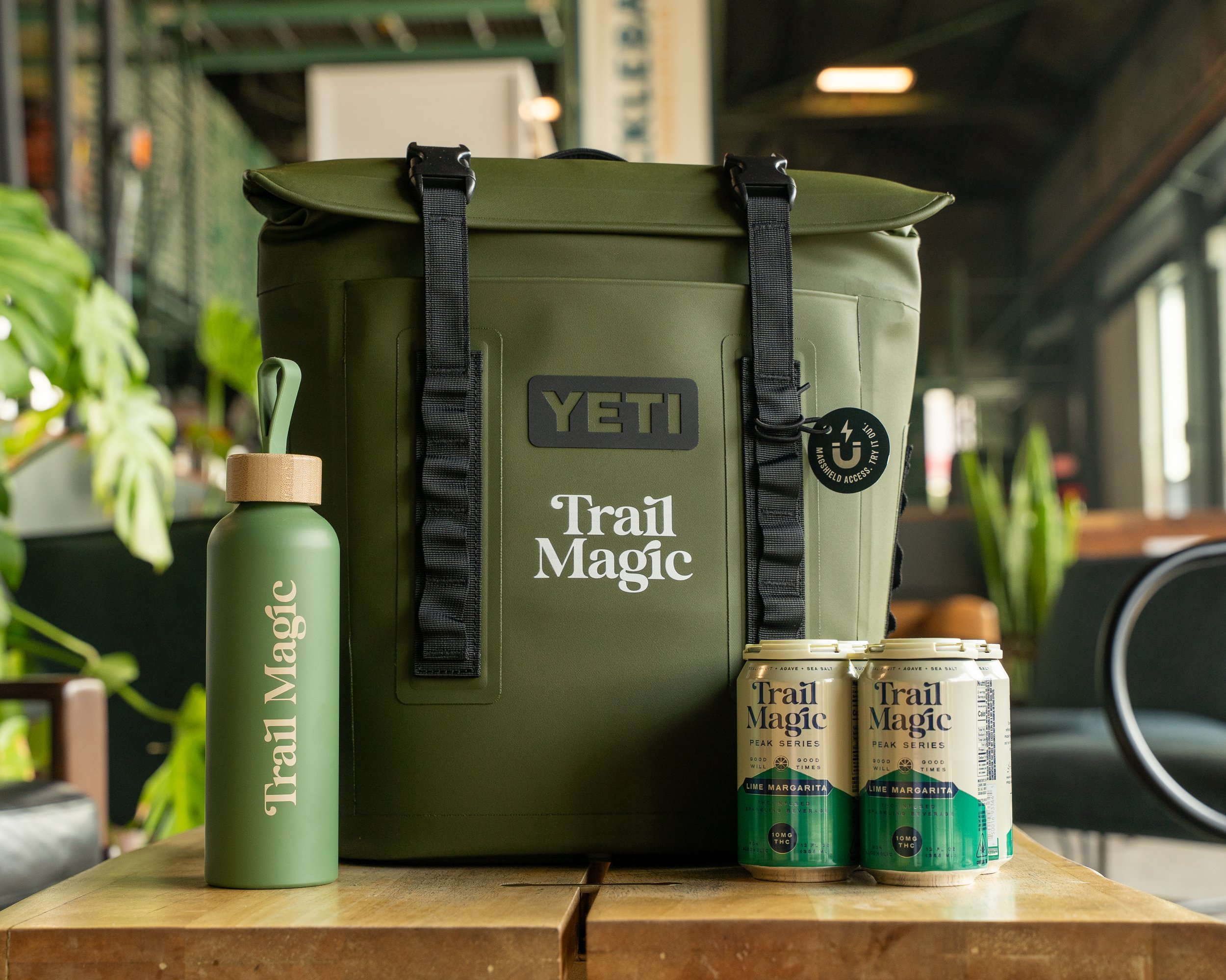 A YETI backpack, a green water bottle, and four cans of Trail Magic Lemon Margarita on a wooden table in a cafe with plants and seating in the background.