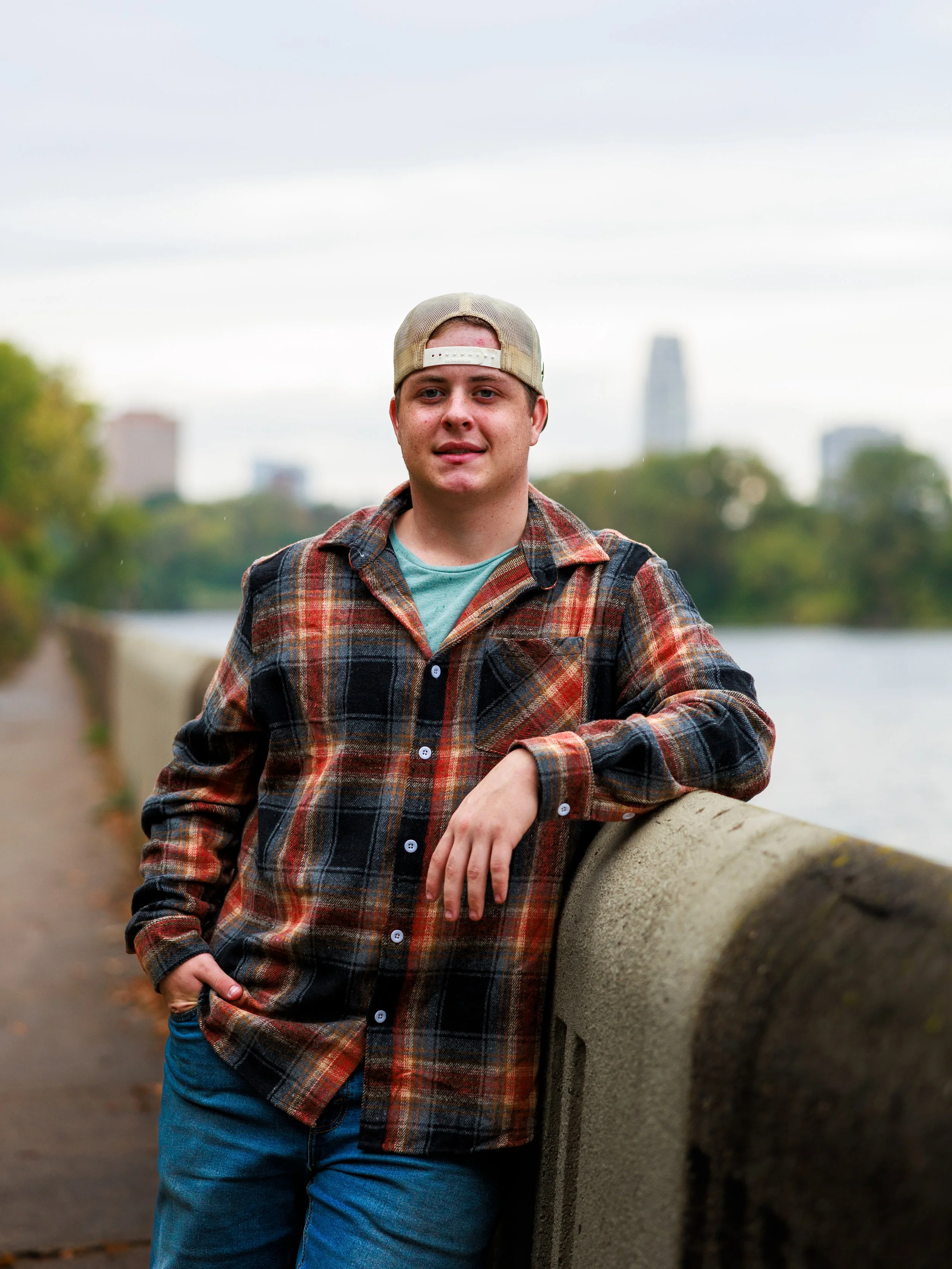 A young man wearing a flannel shirt and a backwards baseball cap leaning on a concrete railing by the water with city buildings in the background.