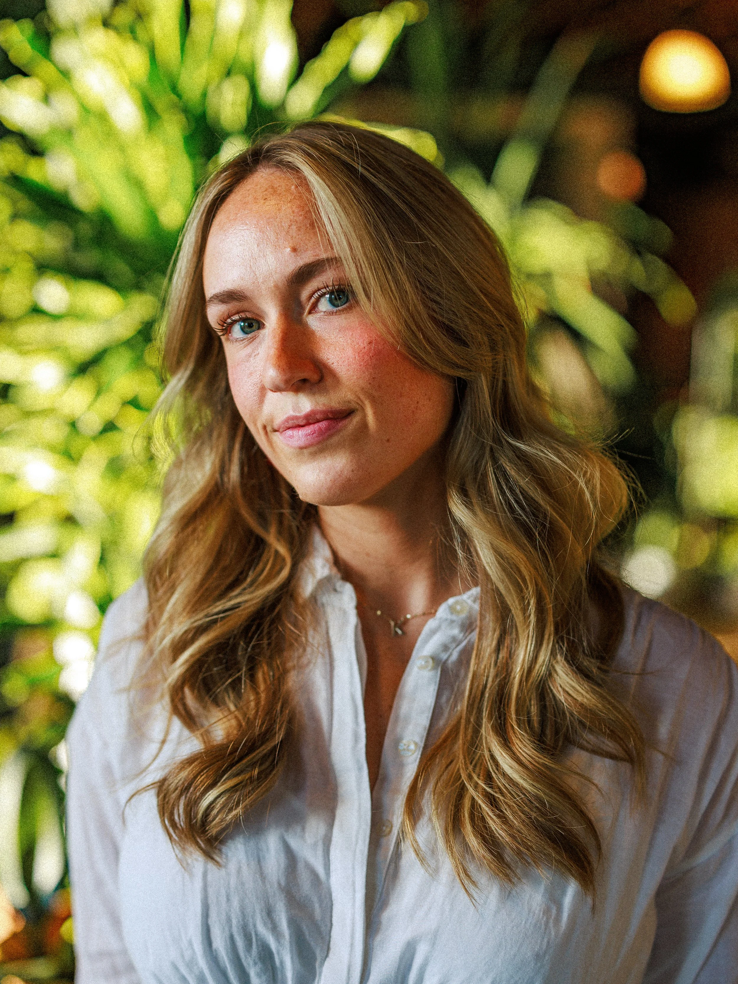 A woman with long, wavy blonde hair and blue eyes, wearing a white button-up shirt and a small necklace, standing outdoors with green foliage in the background.