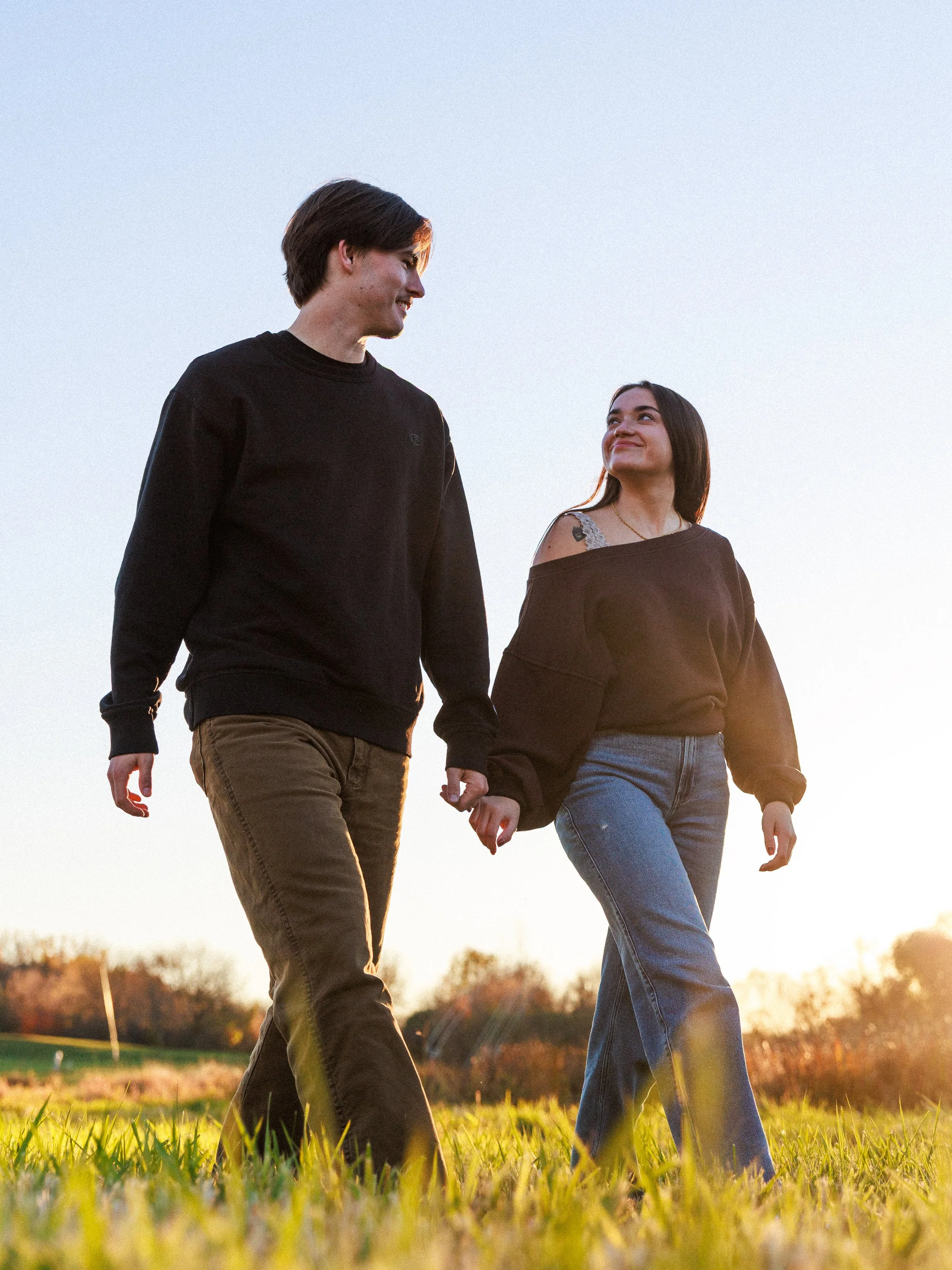 A young couple walking hand-in-hand outdoors during sunset, smiling at each other.