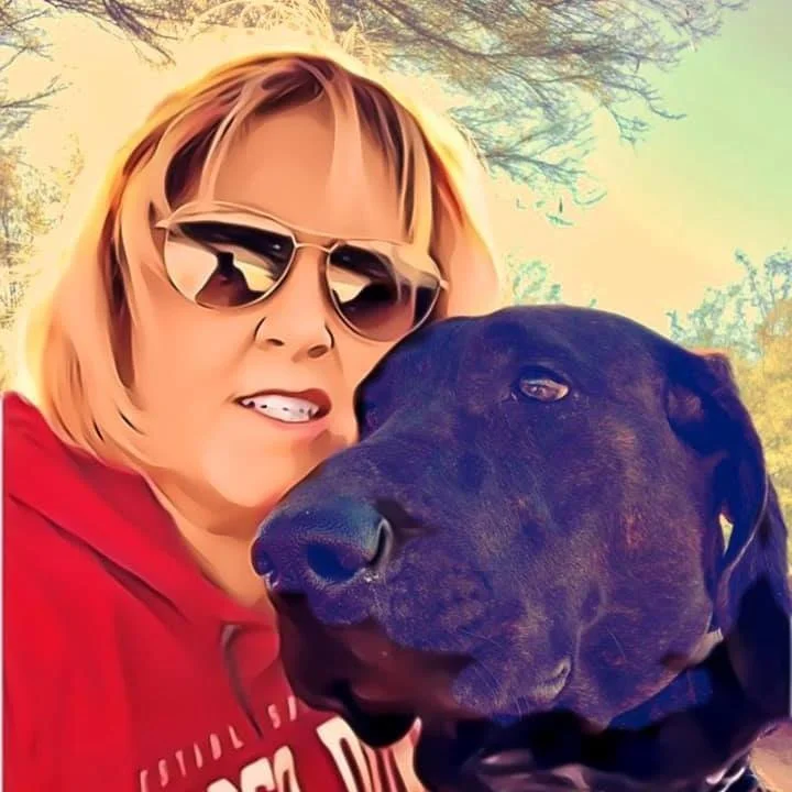 A woman wearing sunglasses and a red hoodie taking a selfie with her black dog outdoors on a sunny day.