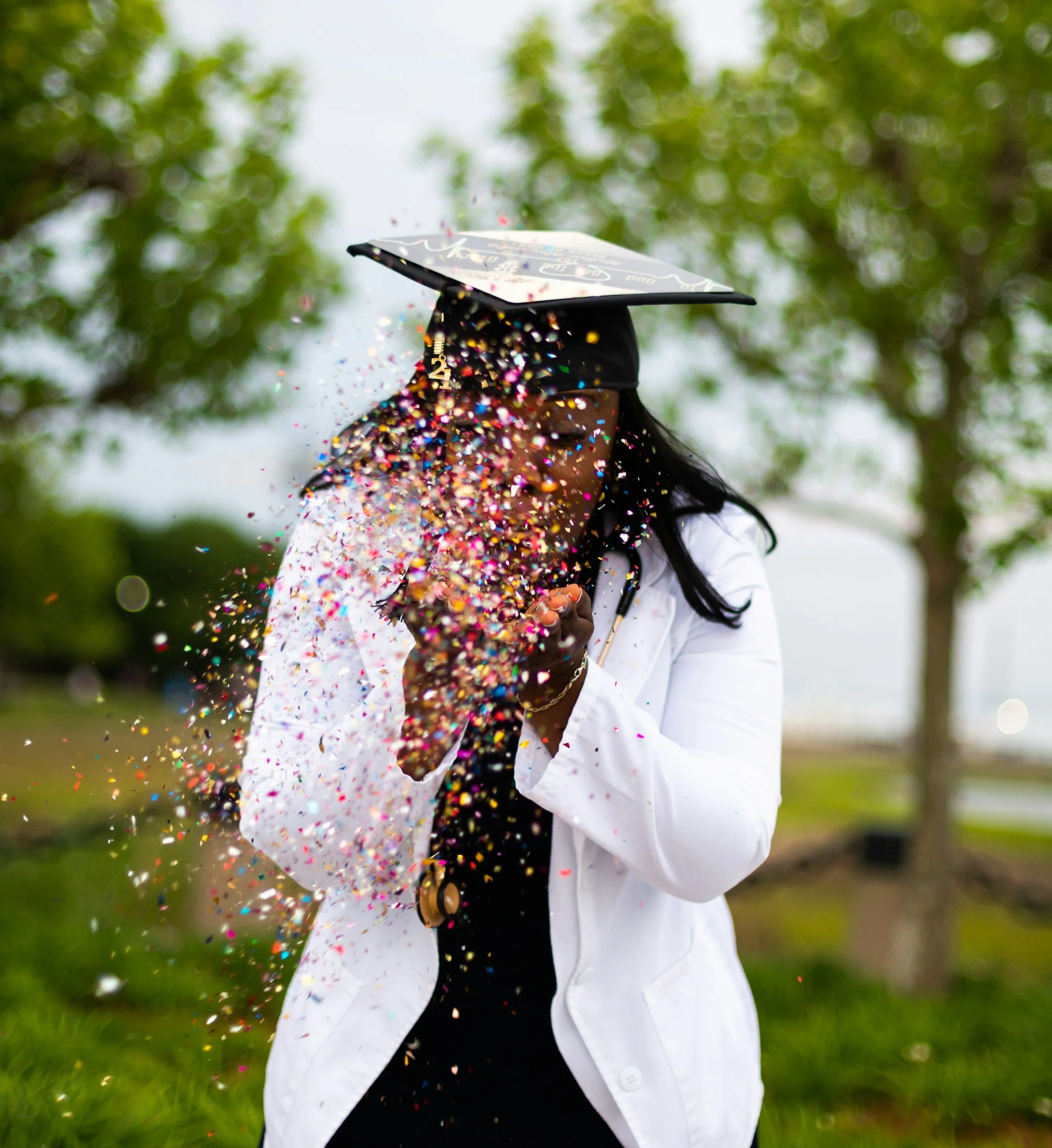 A woman in a white coat and graduation cap celebrating with colorful confetti in an outdoor setting with trees.