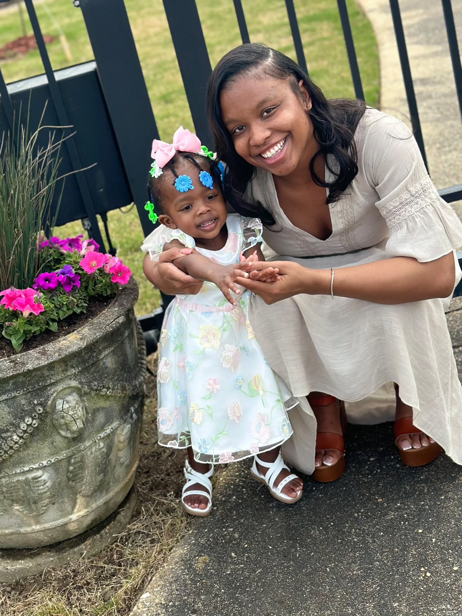 A woman and a young girl smiling and holding hands outdoors near a black metal fence and pink and purple flowers in a large concrete planter.