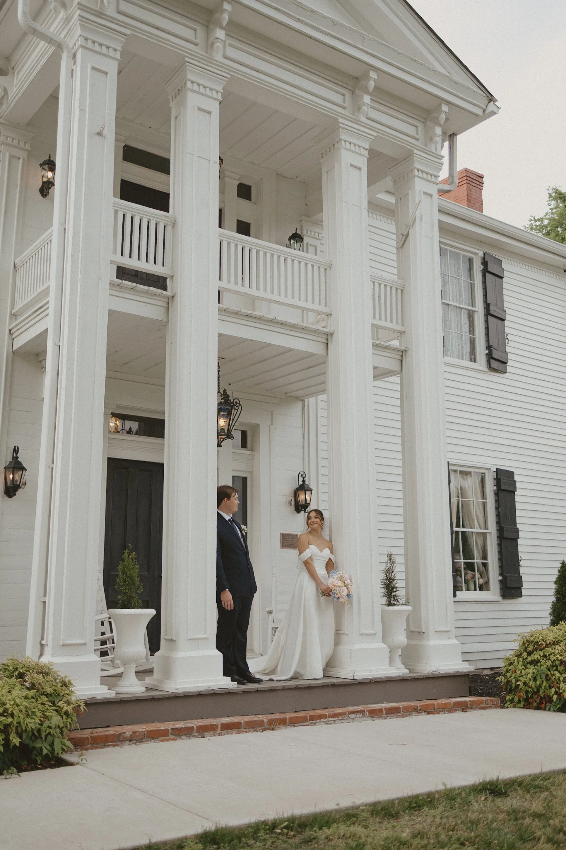 A wedding couple standing outside a white, two-story house with fashionably dark shutters, on a porch with white columns. The bride is wearing a white off-the-shoulder wedding dress holding a bouquet, and the groom is dressed in a black suit, white shirt, and tie.
