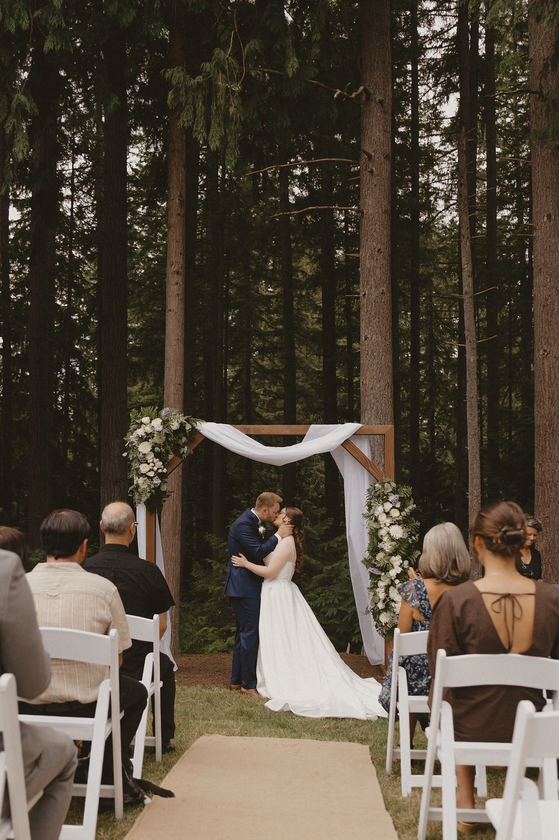 A bride and groom share a kiss during their outdoor wedding ceremony in a forest, with guests seated around them and a decorated wooden arch behind them.