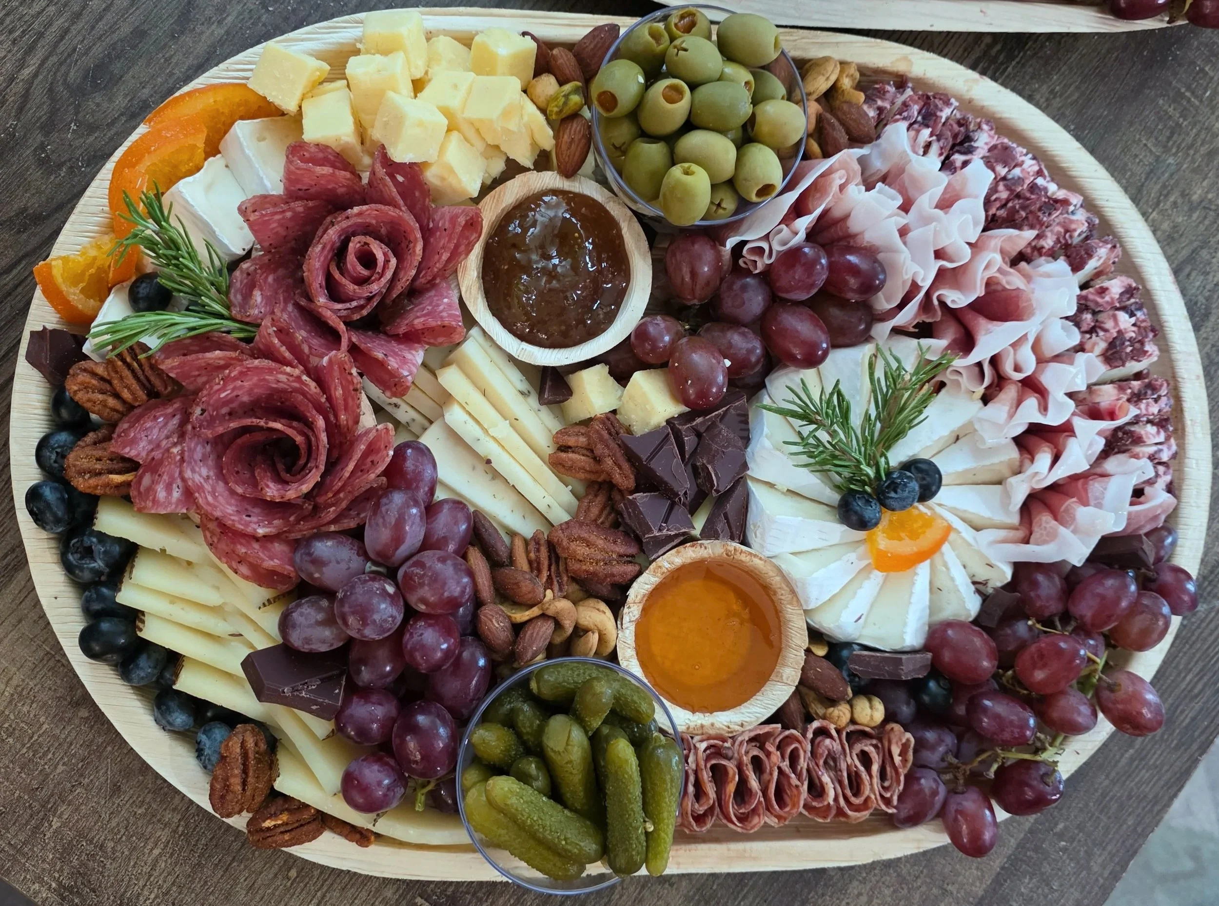 Round wooden charcuterie board with cheese cubes, sliced cured meats, green olives, red grapes, blueberries, almonds, pecans, dark chocolate, honey, and garnished with sprigs of rosemary and slices of orange.