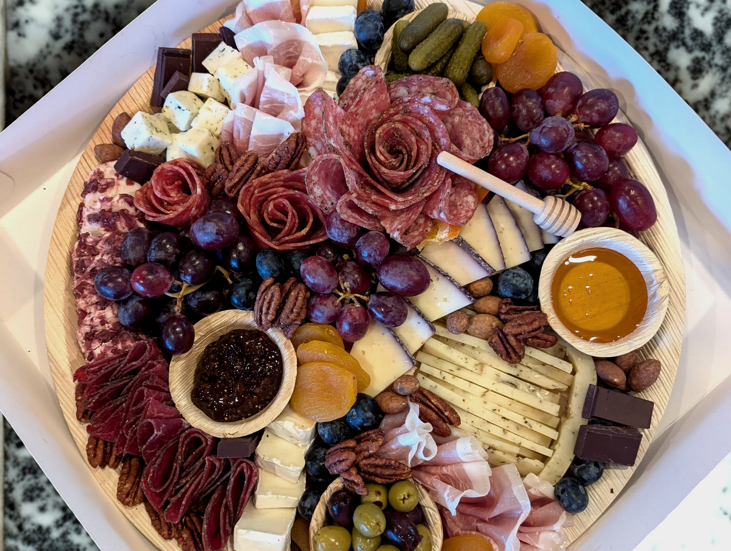 A large charcuterie board with various cheeses, cured meats, grapes, pickles, fruits, nuts, dark chocolate, honey, and jam.