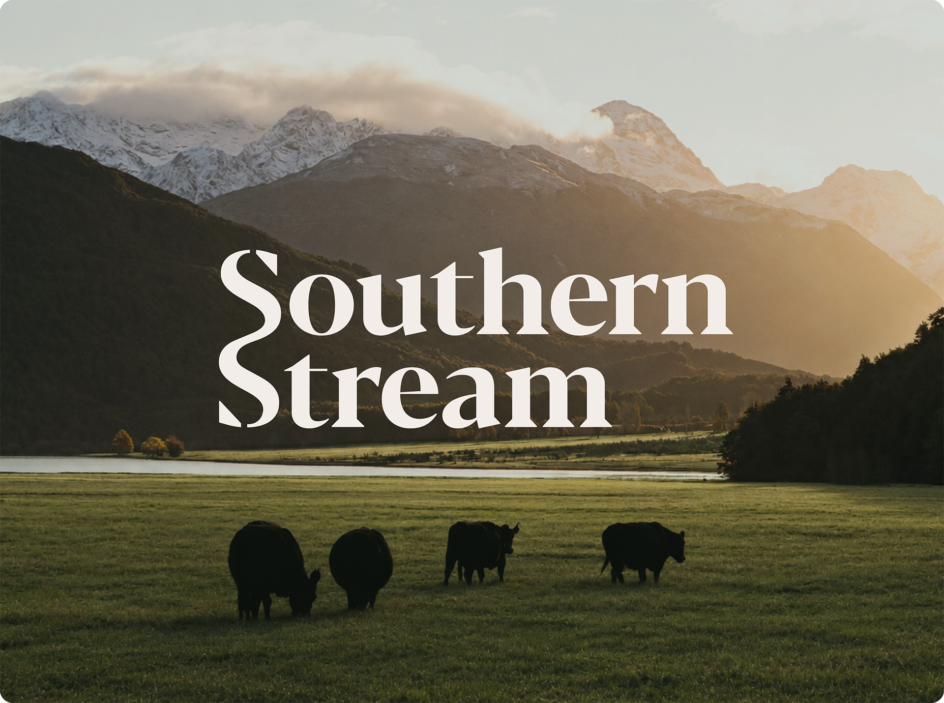 A scenic landscape of mountains with snow and lush green valleys during sunset. Silhouettes of cows grazing in a field in the foreground, with a body of water and trees near the water's edge.