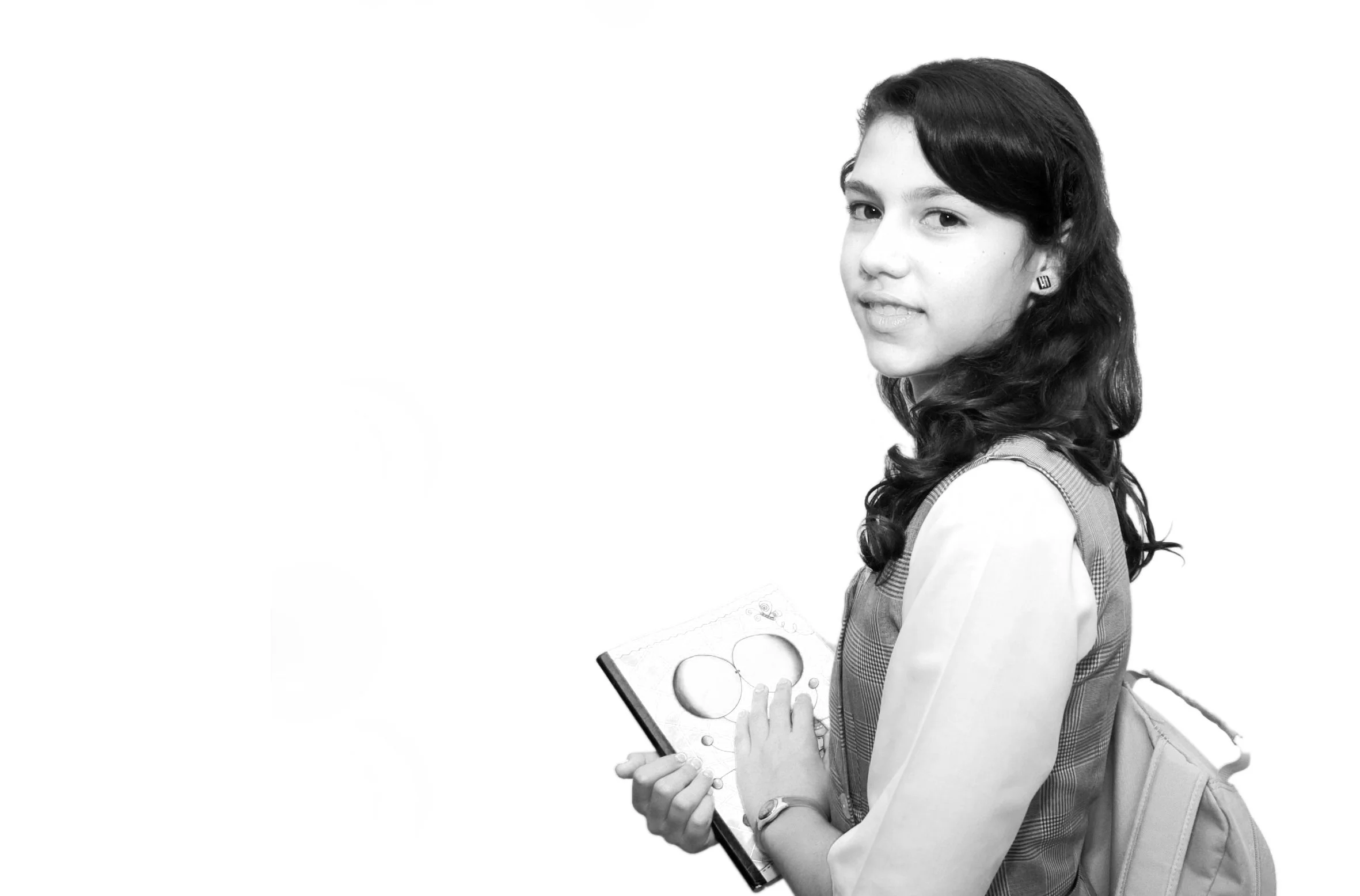 Black and white photo of a schoolgirl with dark wavy hair, wearing a uniform, carrying a backpack, and holding a clipboard with geometric shapes on it, standing and looking at the camera.