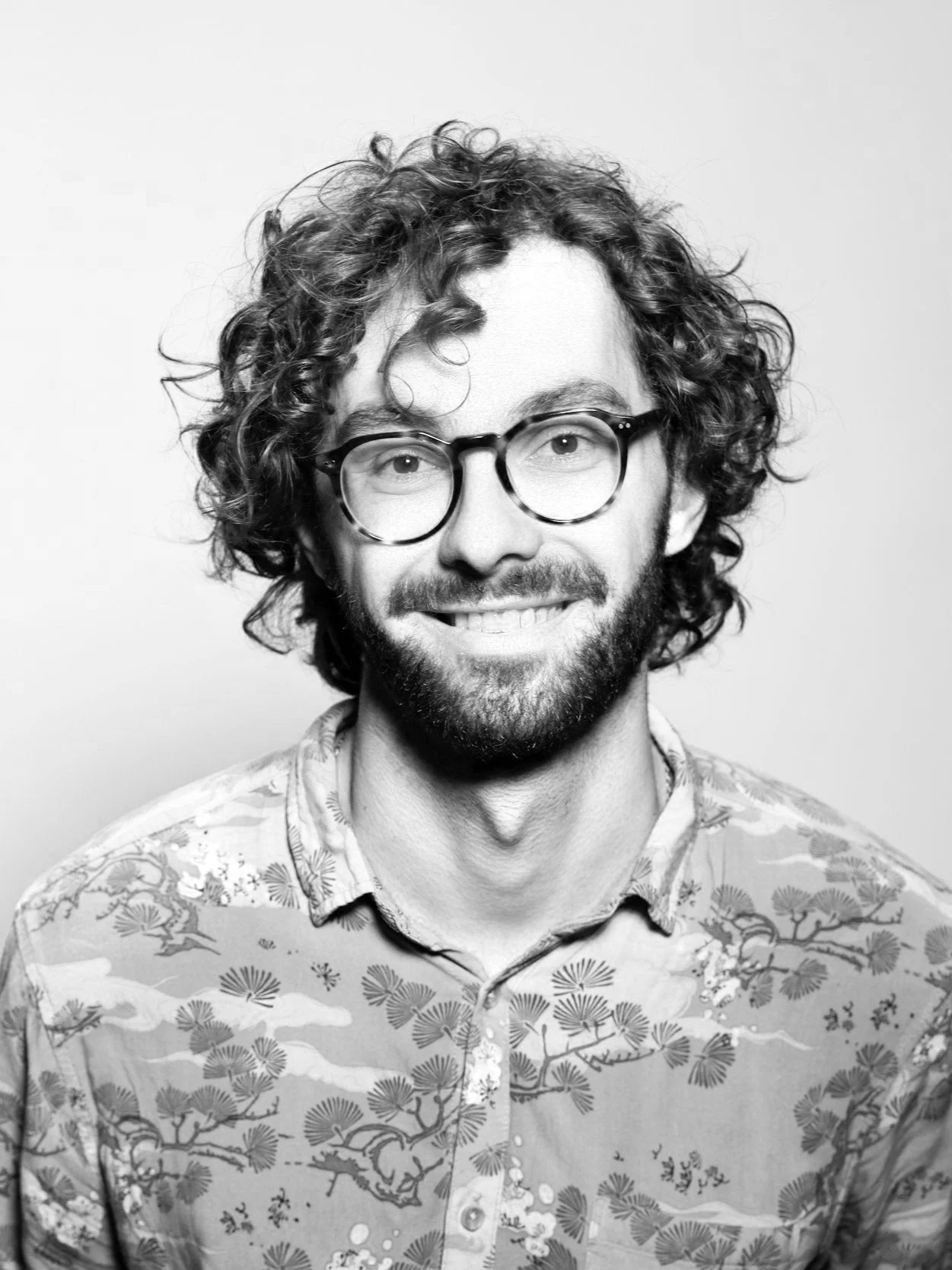 Black and white headshot of a man with curly hair, glasses, beard, wearing a patterned shirt with floral and nature motifs, smiling against a plain background.