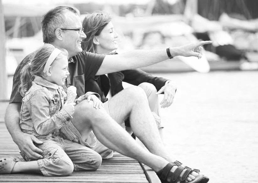 A family of three, including a man, woman, and young girl, sitting on a dock by the water, smiling and enjoying the view of boats in the background. The man is pointing at something in the distance.
