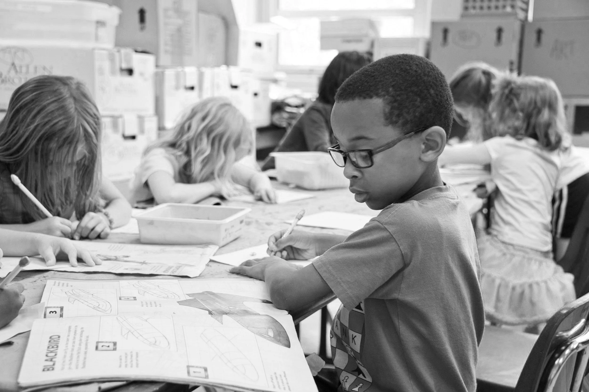 A group of children sitting at a table in a classroom, reading and working on activities from books and papers. One boy in glasses is focused on his work, while others are engaged in their tasks.