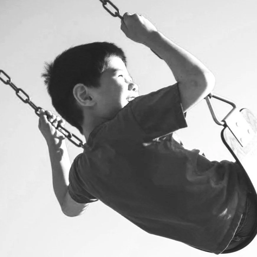 A young boy sitting on a swing, smiling, holding the chains with both hands, looking to the side, in a black and white photo.