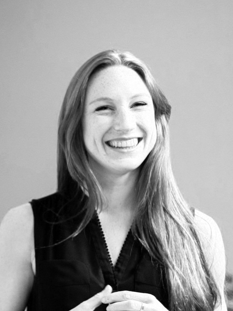 A woman with long hair smiling, wearing a sleeveless top, against a plain background.