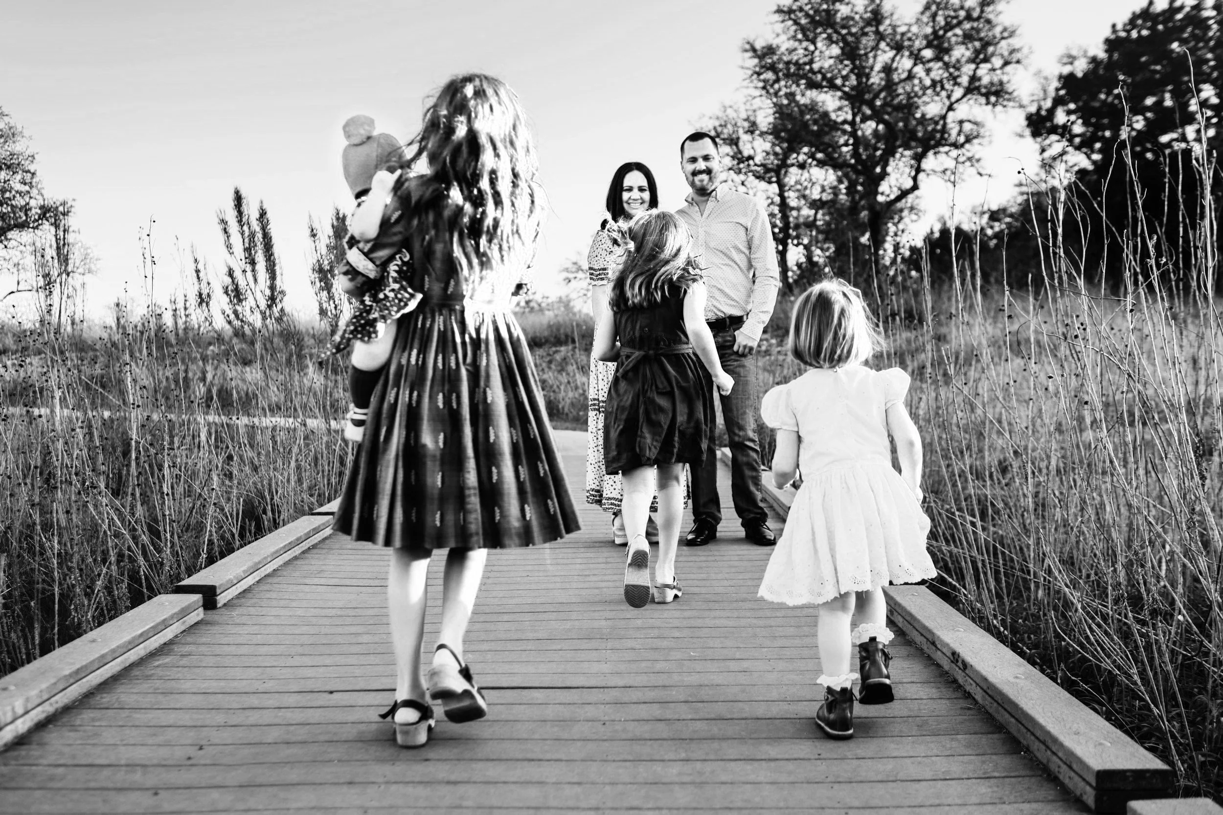 A family walking on a wooden bridge outdoors, with children in the foreground and adults in the background, in a natural setting with trees.