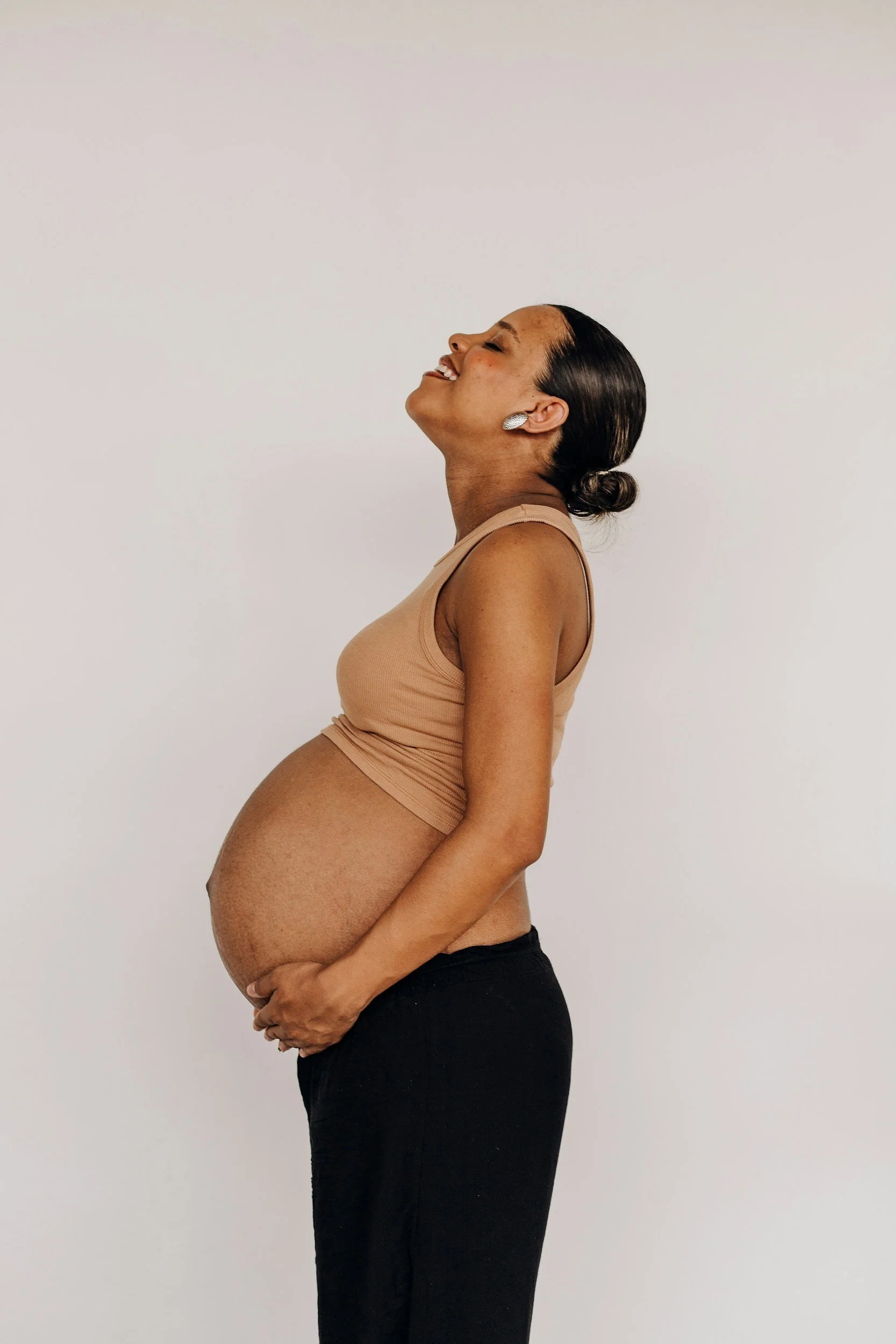 Pregnant woman with dark hair tied back, wearing a beige sleeveless top and black pants, smiling with eyes closed, standing sideways against a plain white wall.