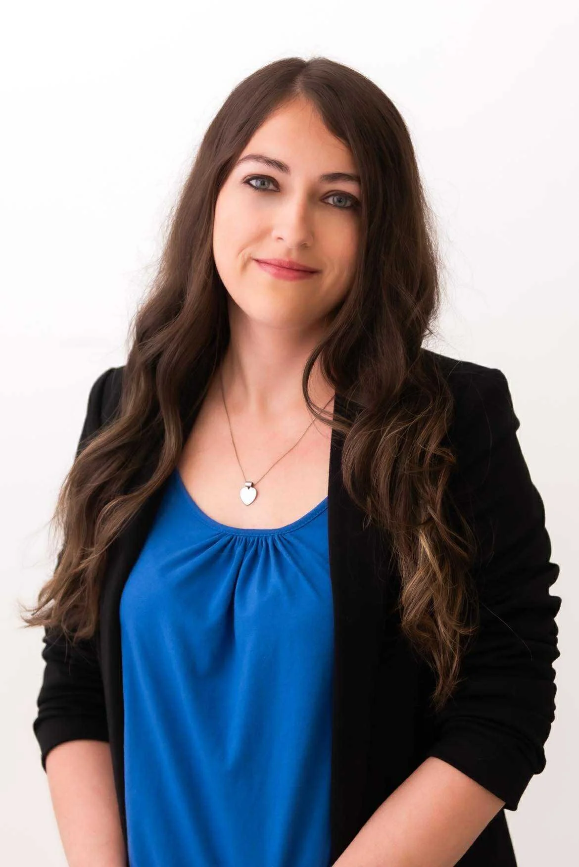 A woman with long wavy brown hair wearing a black blazer and a blue top, smiling softly, standing against a white background.