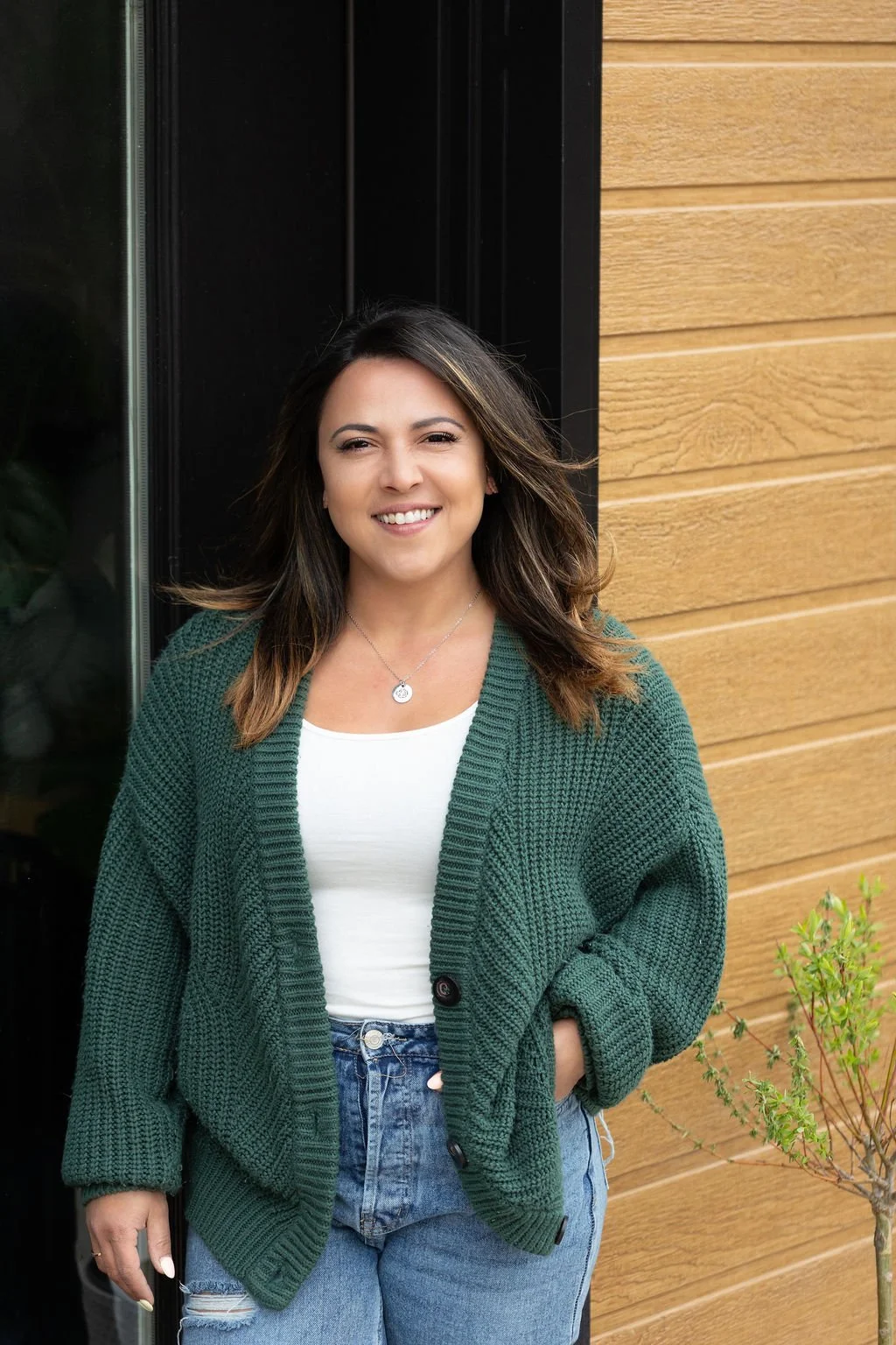 A smiling woman with brown hair wearing a green cardigan and light blue jeans, standing outdoors next to a wooden wall and black window frame.