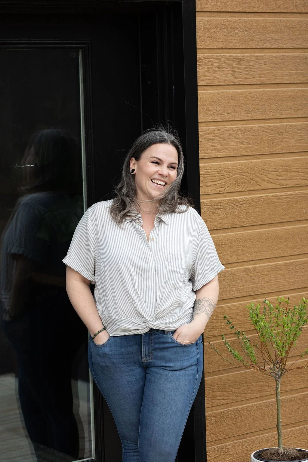 A woman with shoulder-length gray hair, wearing a white striped shirt and blue jeans, is smiling and standing outside near a black door and a small potted tree.