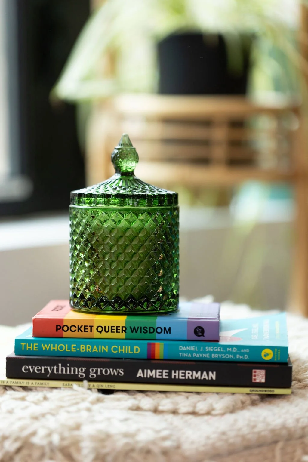 A green glass decorative jar with a lid is placed atop a stack of four books on a cream-colored surface. The books are titled "Pocket Queer Wisdom," "The Whole-Brain Child," "Everything Grows," and "Aimee Herman." In the background, a blurred window and wooden furniture are visible.