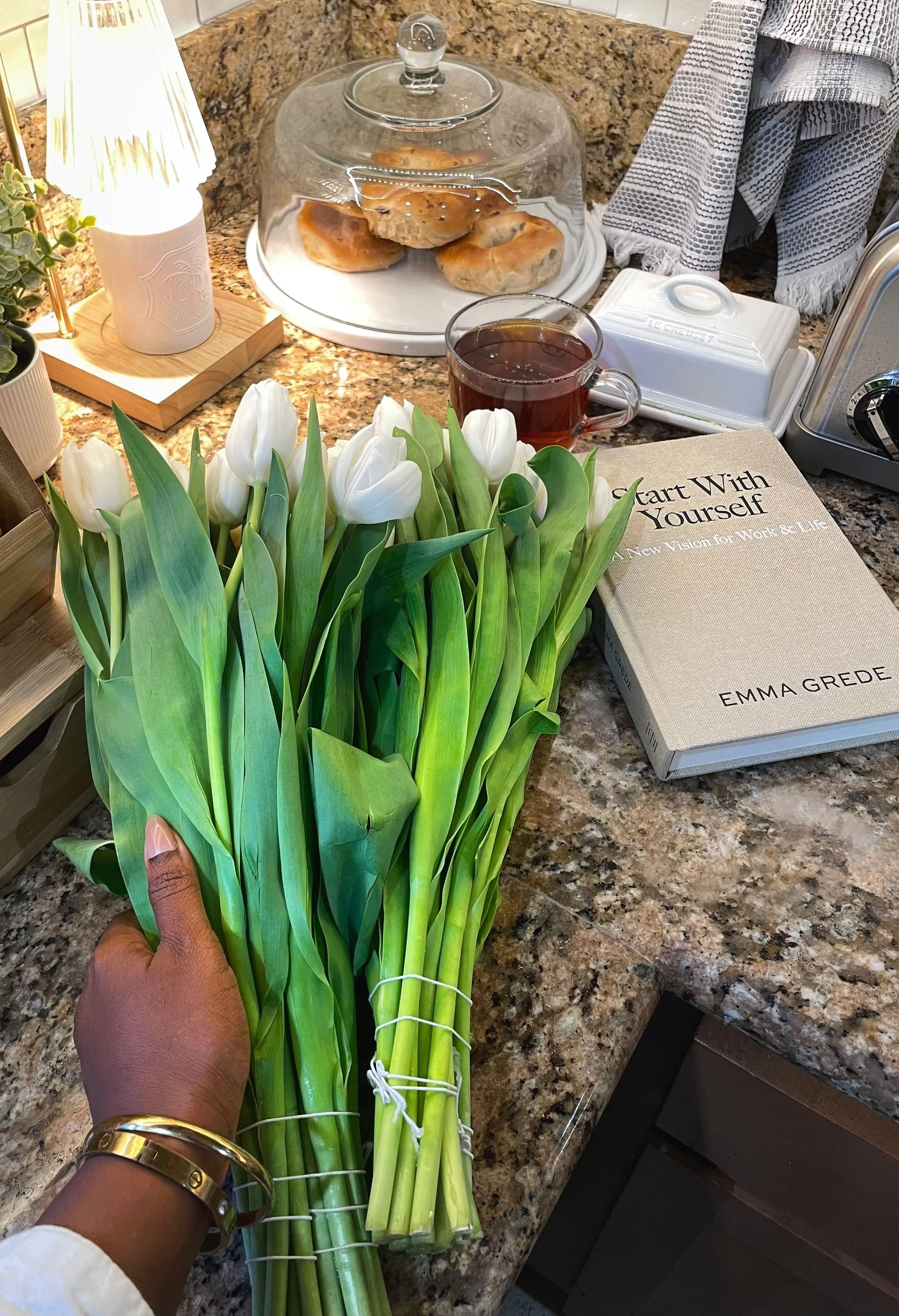 White tulips and Emma Grede Start With Yourself book on kitchen counter with morning tea, coffee, and pastries