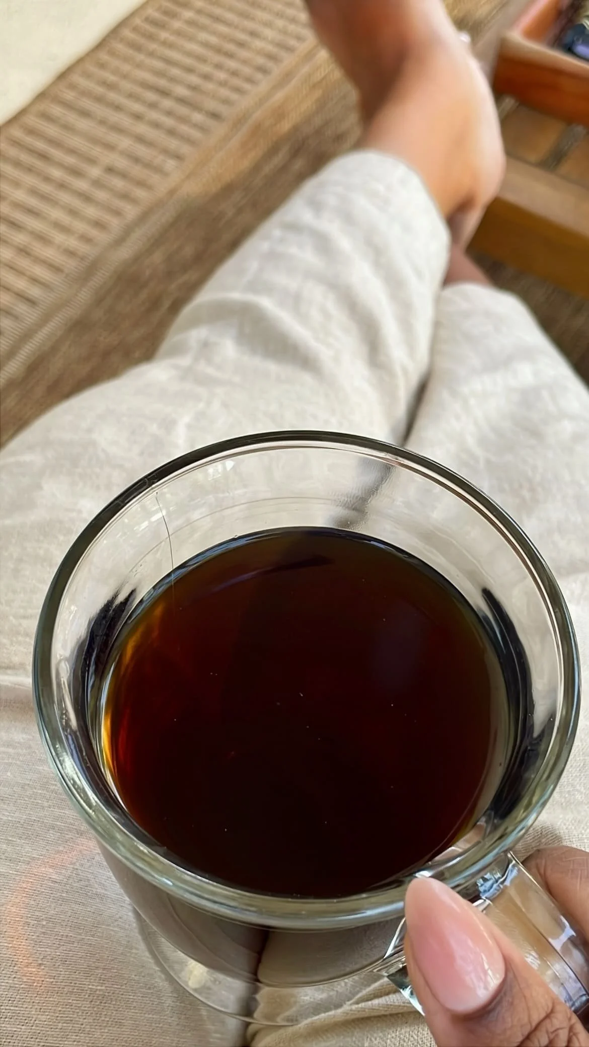 Glass mug of black coffee held in hands while relaxing on balcony in linen pants