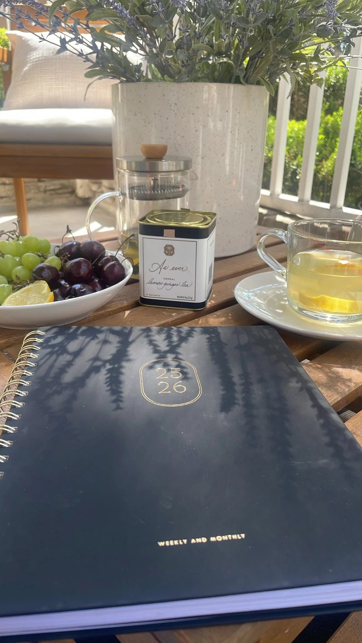 2025–2026 weekly and monthly planner on a wooden patio table beside a glass of As Ever lemon ginger tea, grapes, and a French press in morning sunlight.