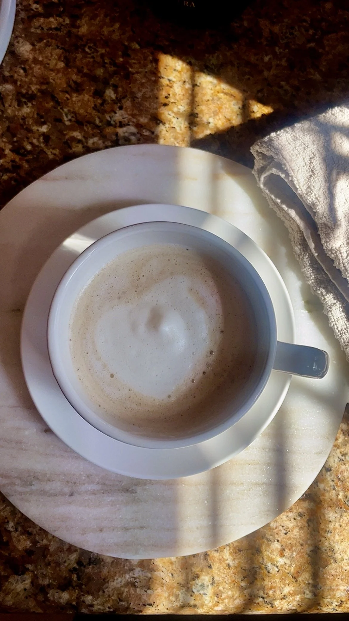 White ceramic cup of coffee on a saucer, resting on a stone countertop with soft morning light and shadows.