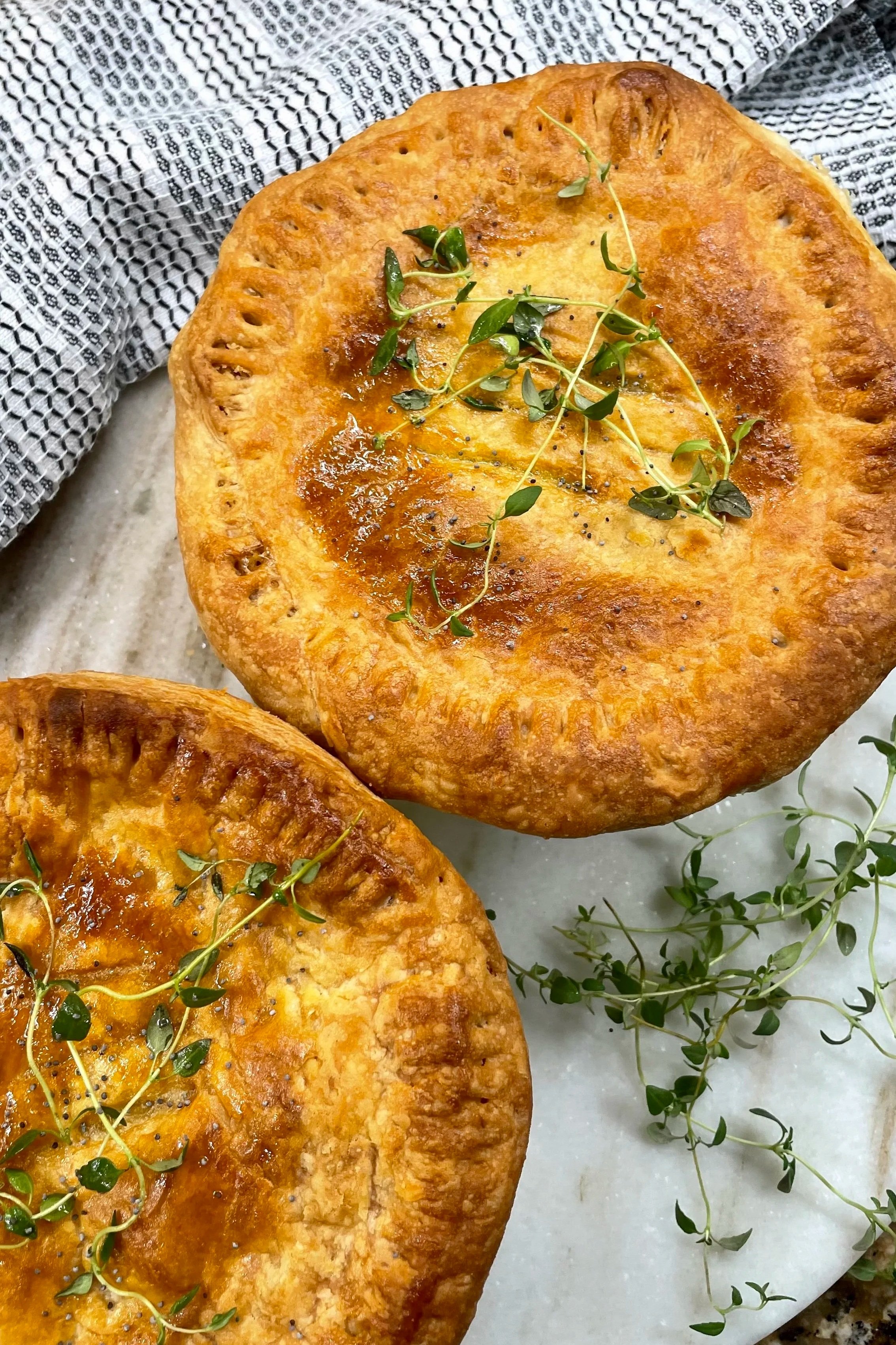 Golden-brown homemade chicken pot pies with flaky puff pastry crusts, garnished with fresh thyme and poppy seeds on a white marble serving board.