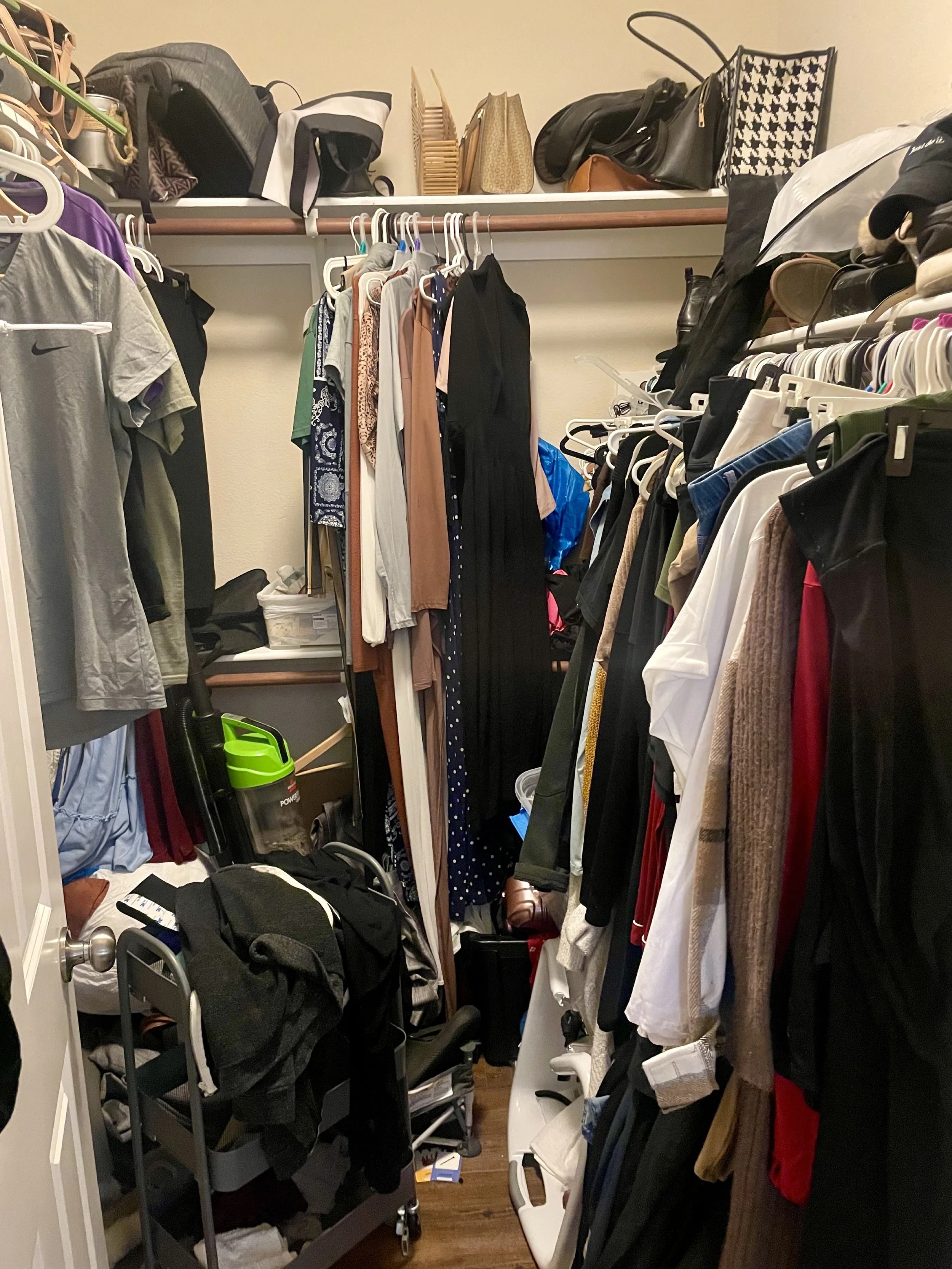 Overcrowded bedroom closet with mixed hangers, cluttered floor, vacuum tucked between clothes, and disorganized shelves contributing to daily stress.