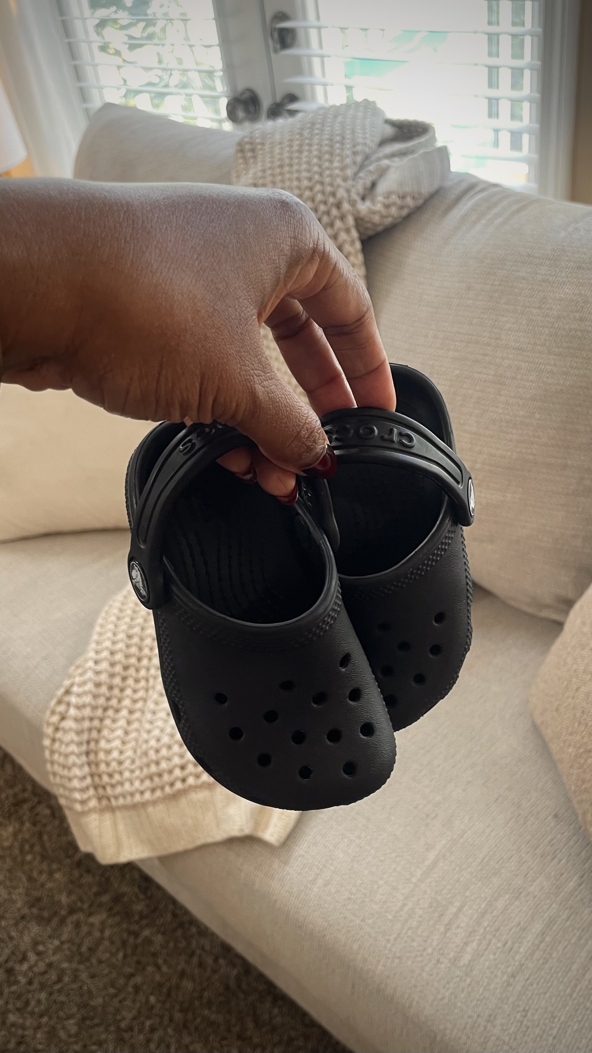 A woman's hand holding a pair of black crocs with a soft beige chenille couch and blanket in the background